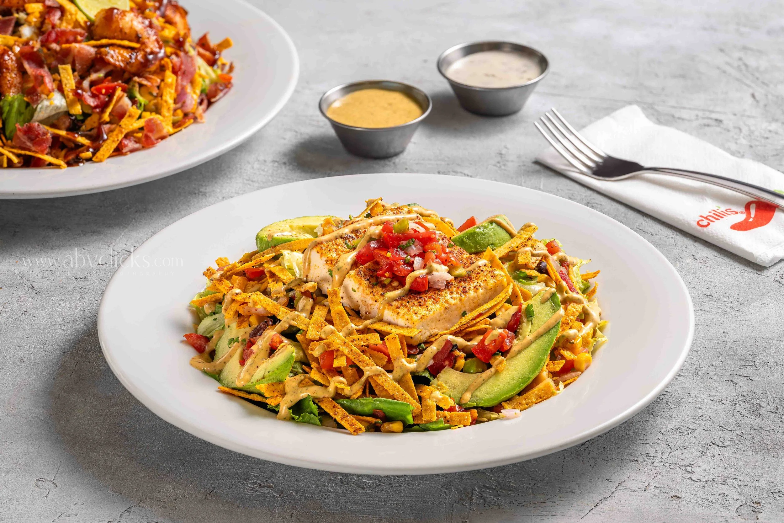 A white plate of grilled chicken topped with diced tomatoes, onions, and sauce, served over sliced avocado, shredded cheese, and crunchy tortilla strips. In the background, another plate of similar dish, along with small containers of creamy and yell