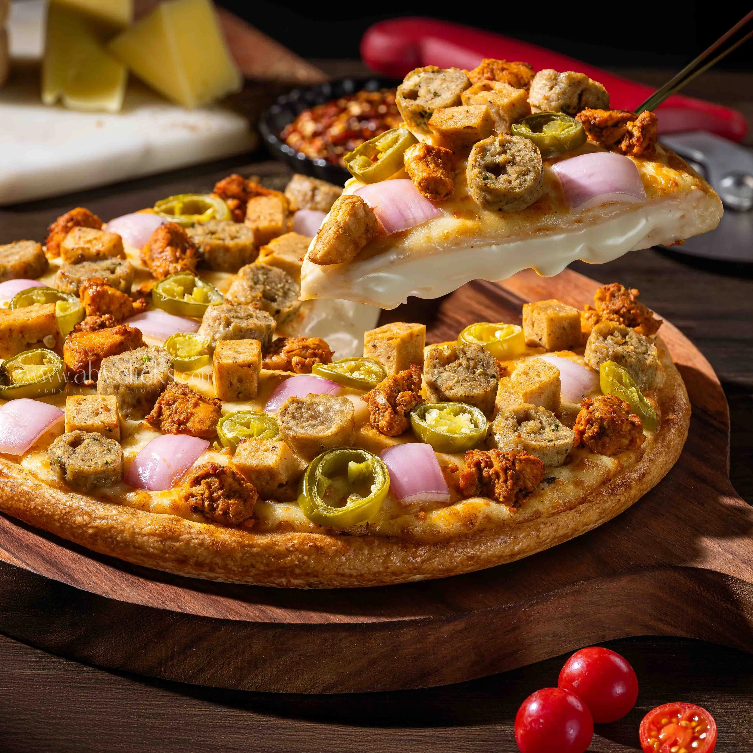 Close-up of a slice of pizza topped with sliced jalapeños, cooked sausage chunks, and sliced onions on a wooden serving board, with whole and halved cherry tomatoes nearby.
