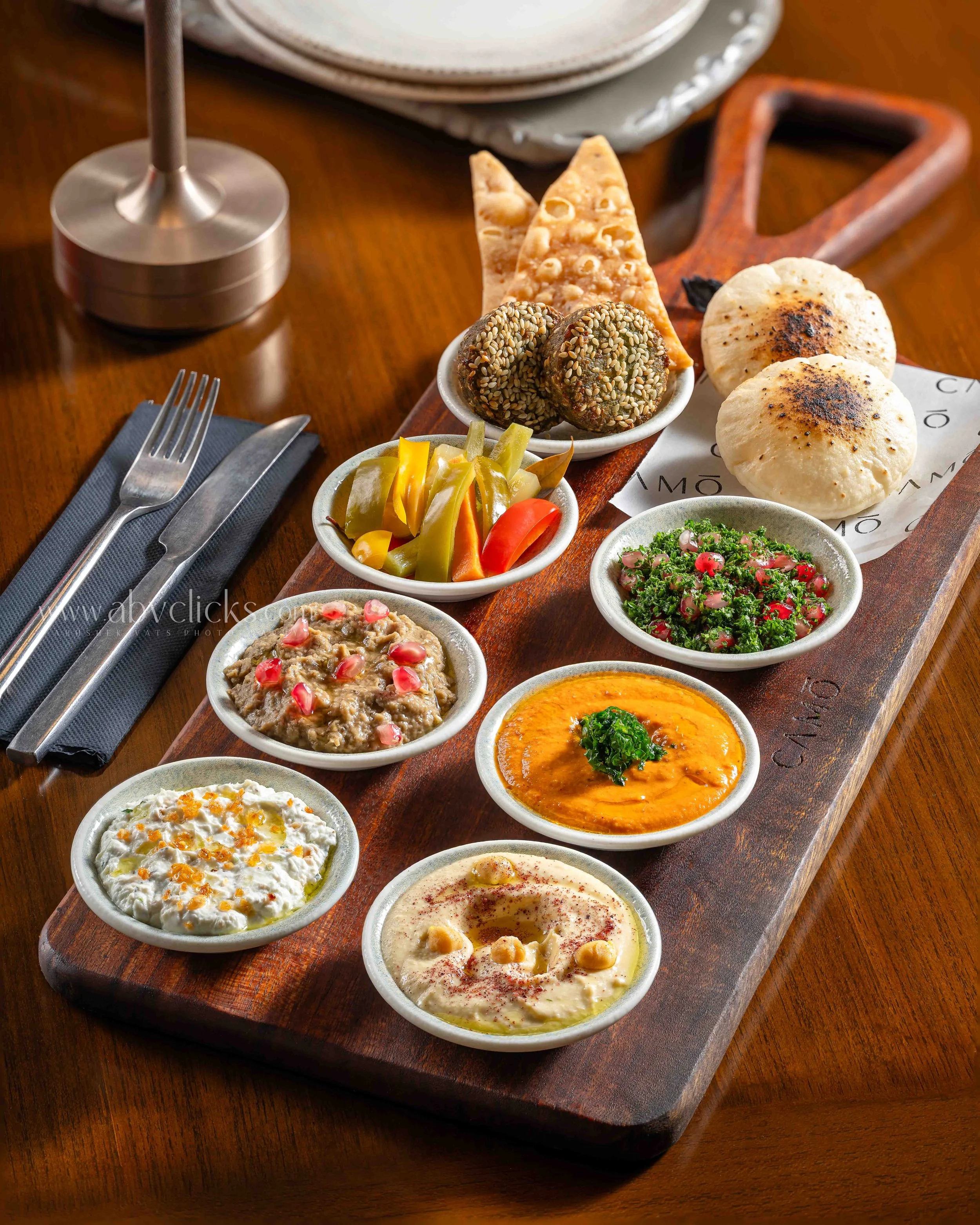 A wooden serving board with small bowls of various Mediterranean and Middle Eastern dips, spreads, and salads, including hummus, labneh, tabbouleh, and baba ganoush, along with pita bread and fresh vegetables on a wooden table.
