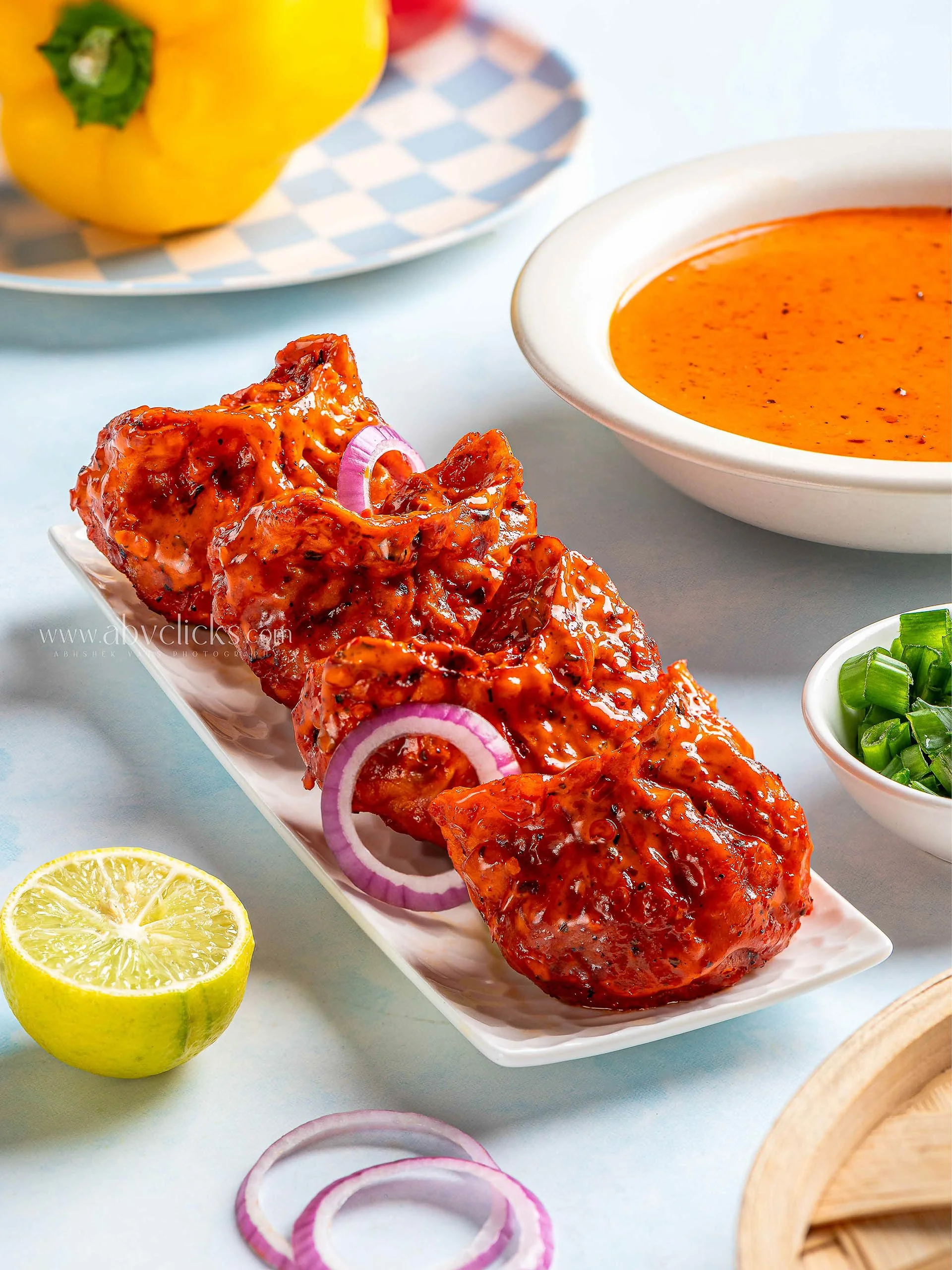 Close-up of tandoori chicken with onion slices, lemon, and side dishes on a white rectangular platter.