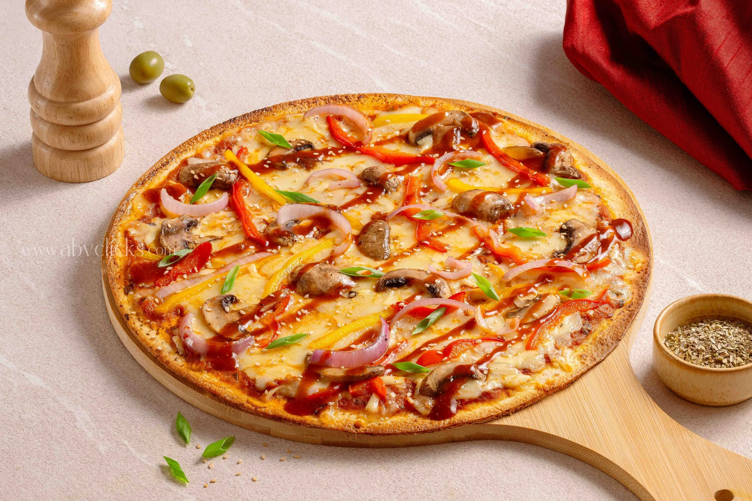 Freshly baked pizza topped with sausage, red and yellow bell peppers, onions, mushrooms, and green herbs, placed on a round wooden board on a light-colored table.