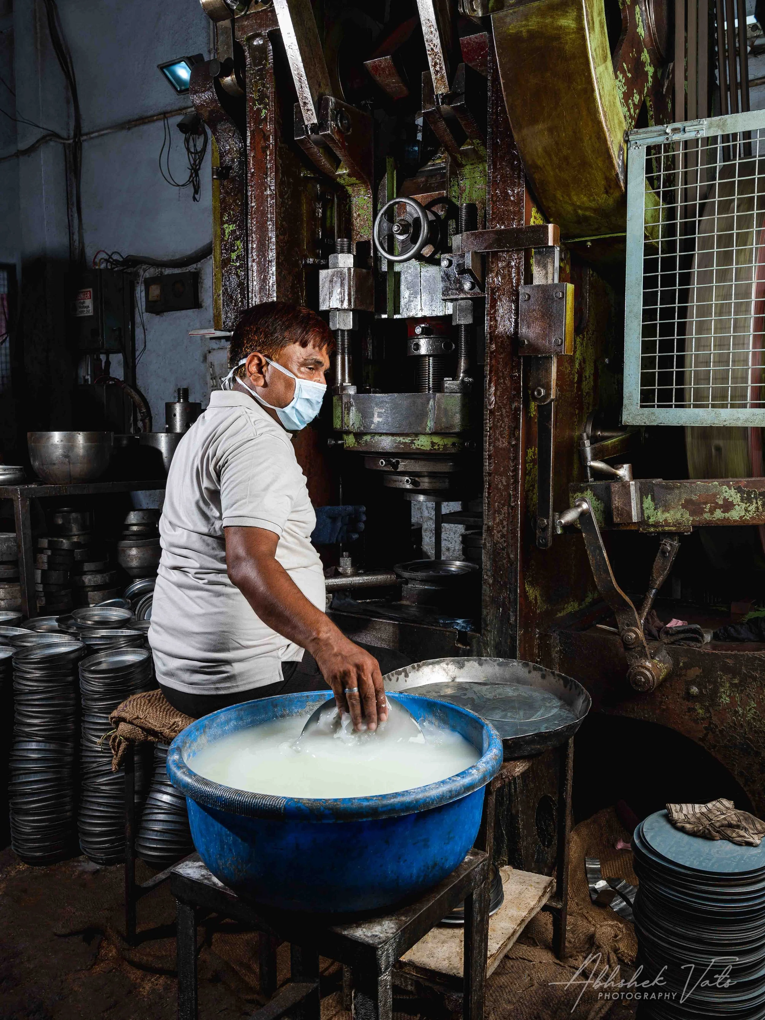 A man wearing a face mask and gloves is working with a large industrial machine, possibly for manufacturing or processing metal or other materials. He is standing beside a large blue container filled with a milky liquid, with additional stacks of bla