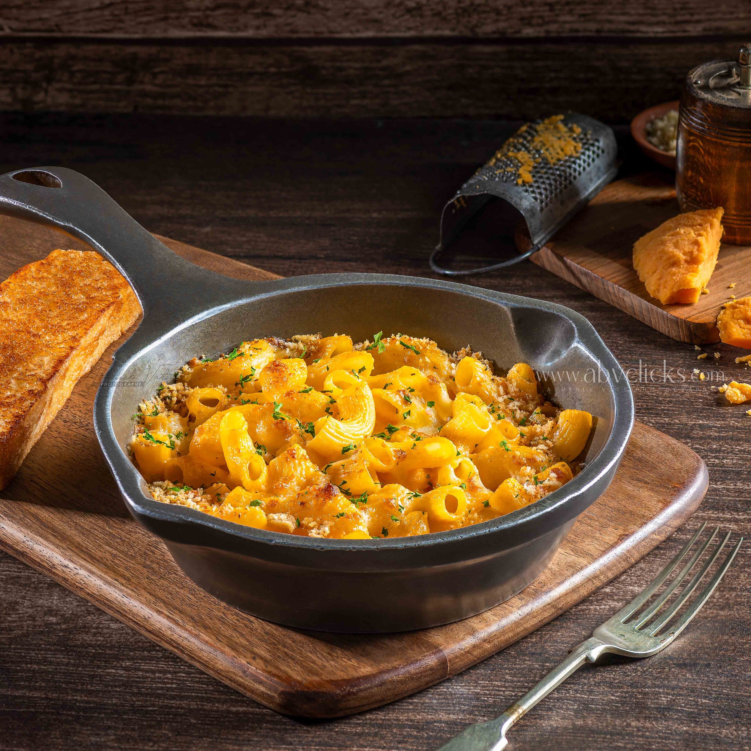 A skillet of baked mac and cheese with a golden crust, topped with parsley, placed on a wooden board with a serving spoon nearby.