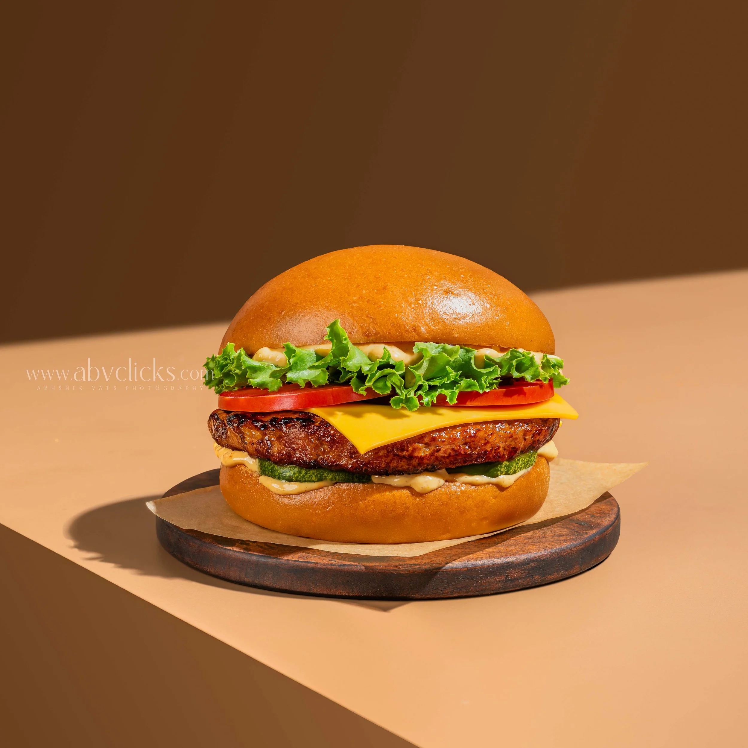 Close-up of a burger with lettuce, tomato, cheese, a beef patty, and a bun, placed on a dark wooden surface against a brown background.