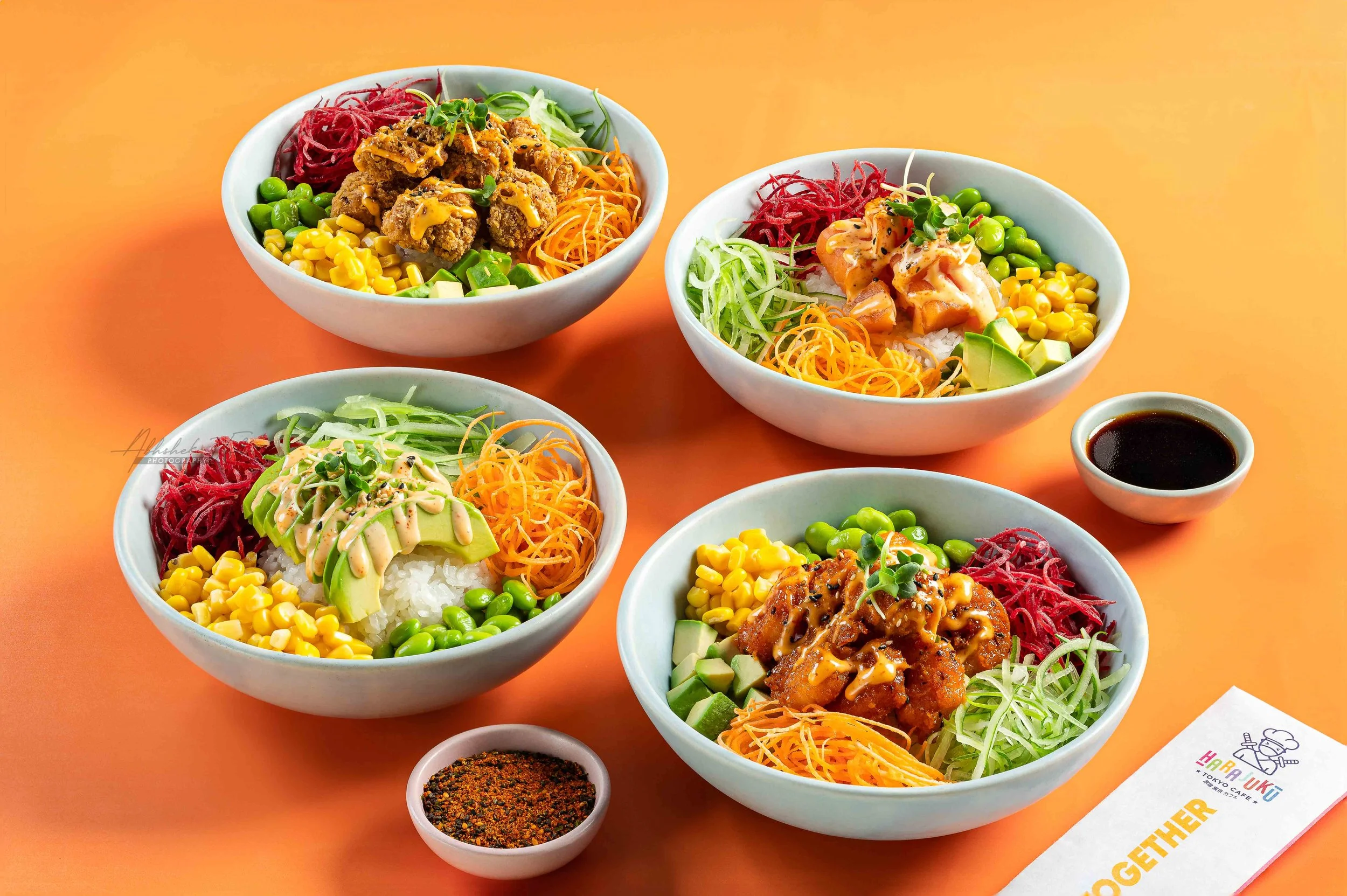 Several bowls of colorful sushi and poke bowls on a bright orange background with small dishes of soy sauce and seasoning.