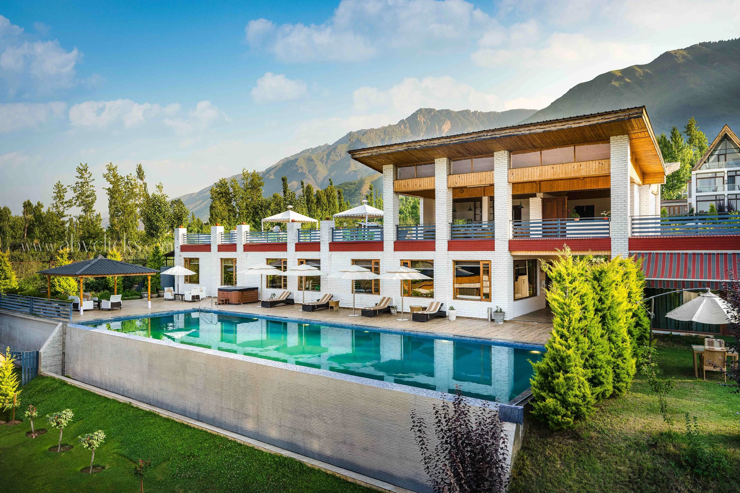A luxurious outdoor swimming pool adjacent to a modern multi-story house with a mountain view in the background. The pool area features lounge chairs, umbrellas, and greenery, under a partly cloudy sky.