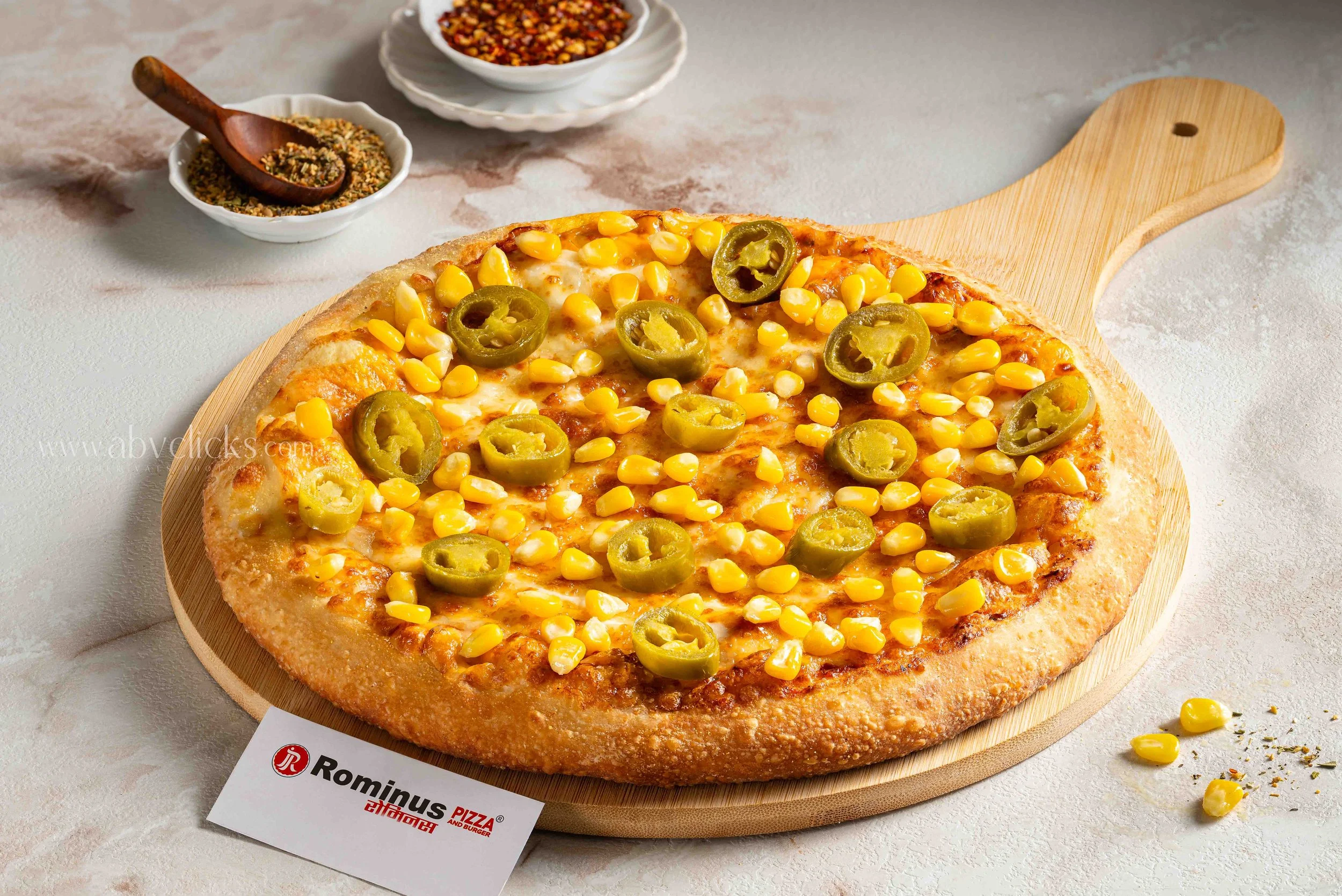 A pizza topped with yellow corn and green jalapeno slices on a wooden pizza board, with small bowls of crushed red pepper flakes and seasoned herbs in the background.