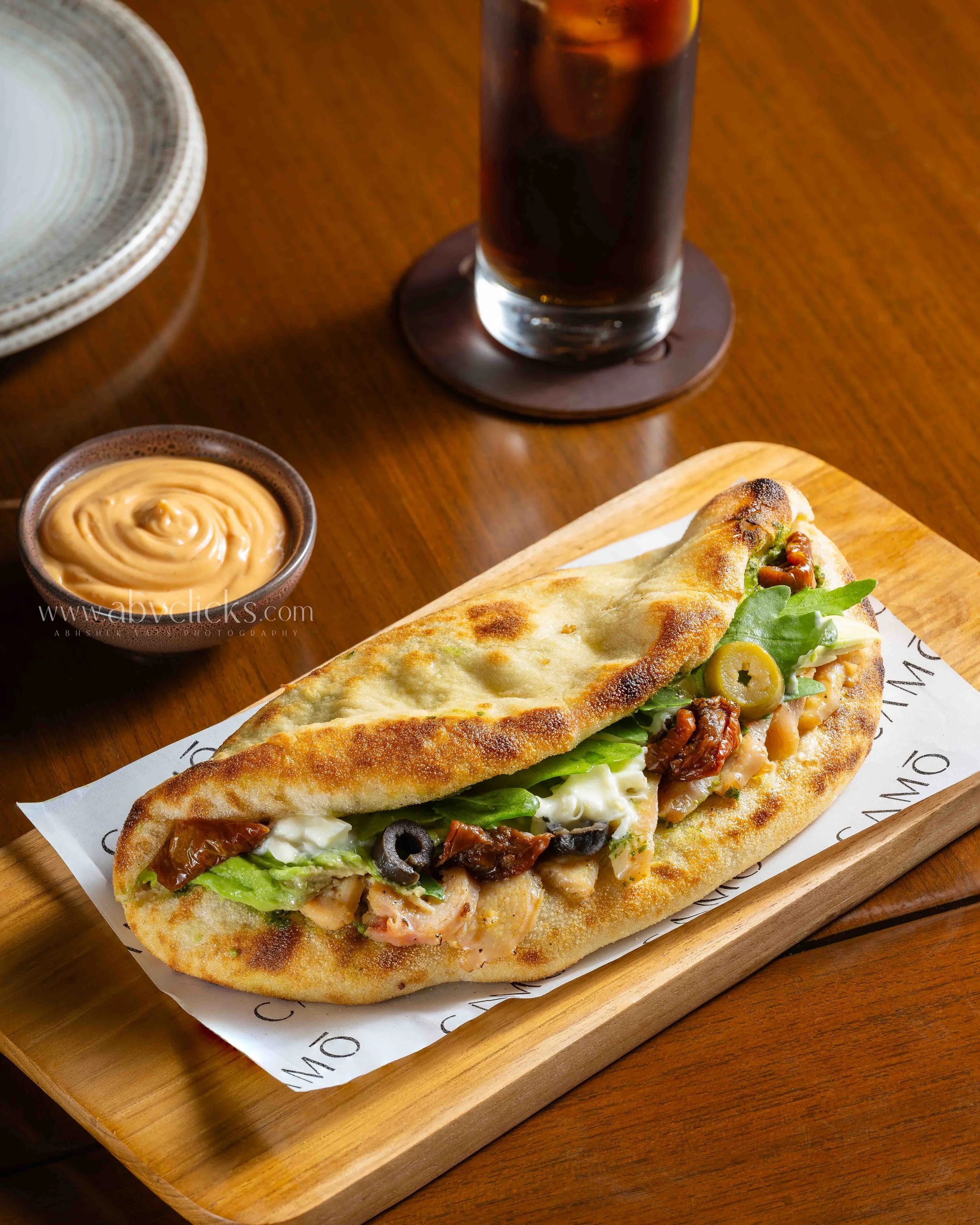 A calzone pizza with toppings including black olives, green olives, sun-dried tomatoes, and fresh greens on a wooden tray, accompanied by a small bowl of dipping sauce and a glass of iced soda.
