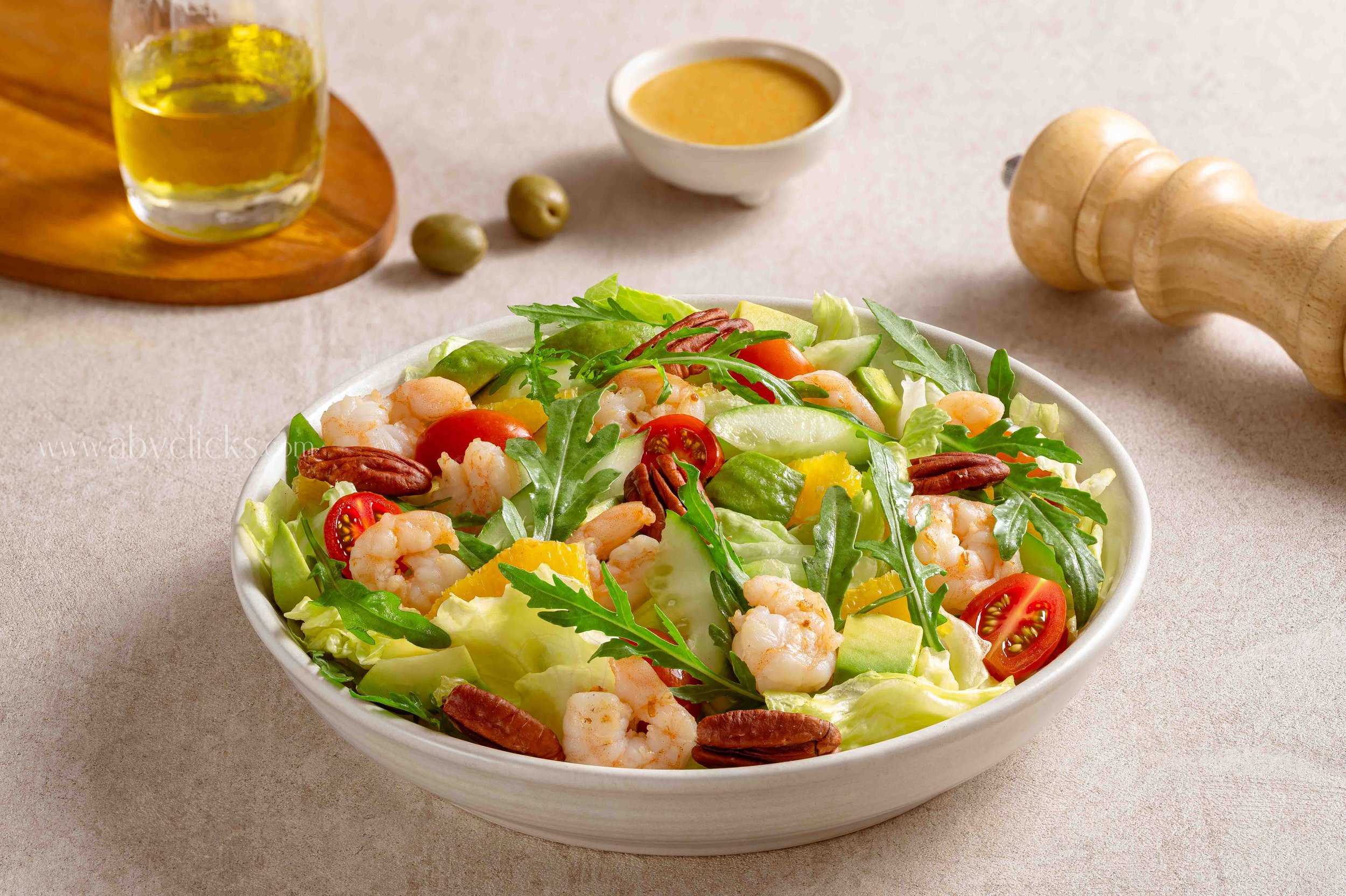 A bowl of mixed salad with shrimp, cherry tomatoes, avocado, pecans, and lettuce on a beige surface, with a glass of olive oil, a small bowl of dressing, a wooden pepper grinder, and two green olives in the background.