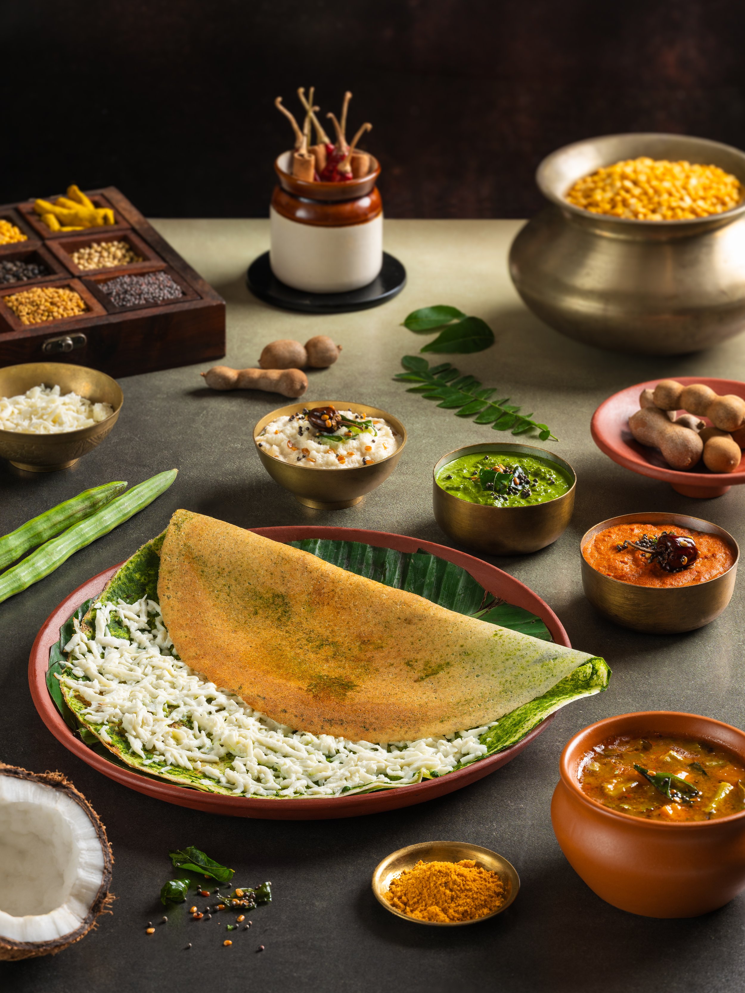 Assorted Indian spices, herbs, chutneys, and dishes including a dosa with coconut chutney, vegetable curry, and sambar, arranged on a dark countertop.