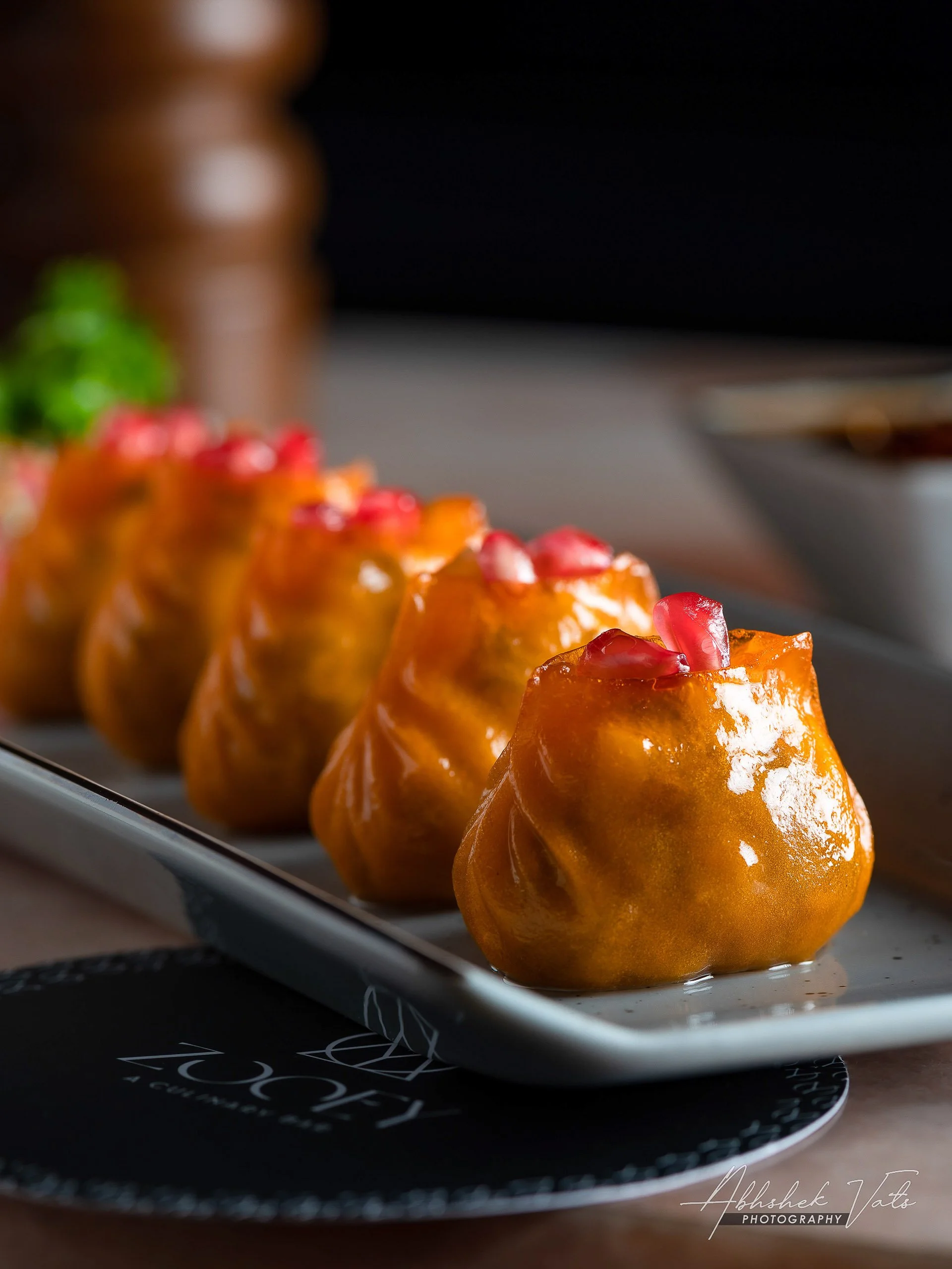 Close-up of several golden brown, glazed Indian sweets topped with pomegranate seeds, arranged on a black plate with modern design, with a blurred dark background.