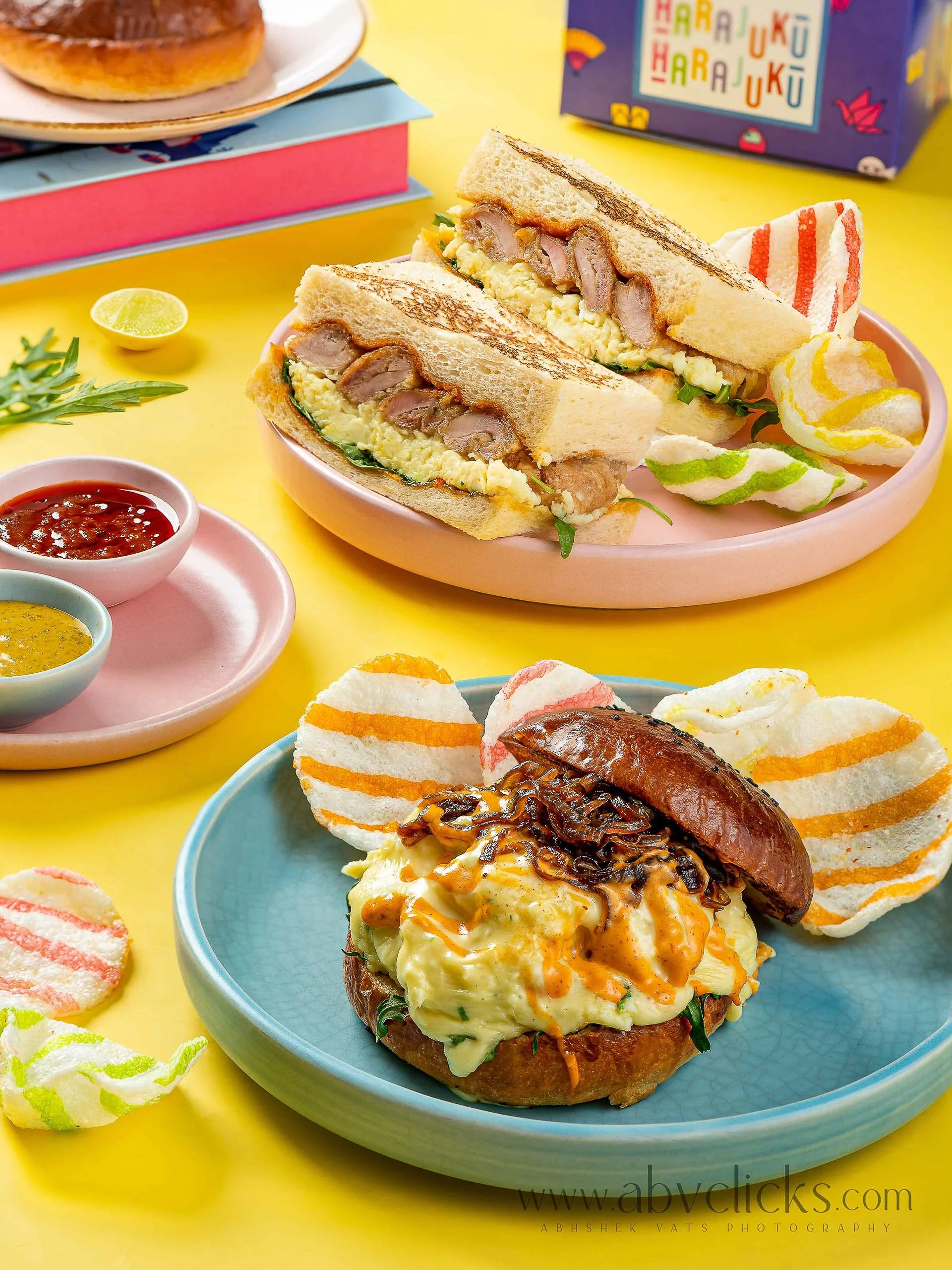 A plate with a burger topped with an egg omelet, caramelized onions, and greens, served with colorful snack chips on the side. In the background, there are small bowls of sauces and a colorful box with the words 'Harajuku' printed on it.