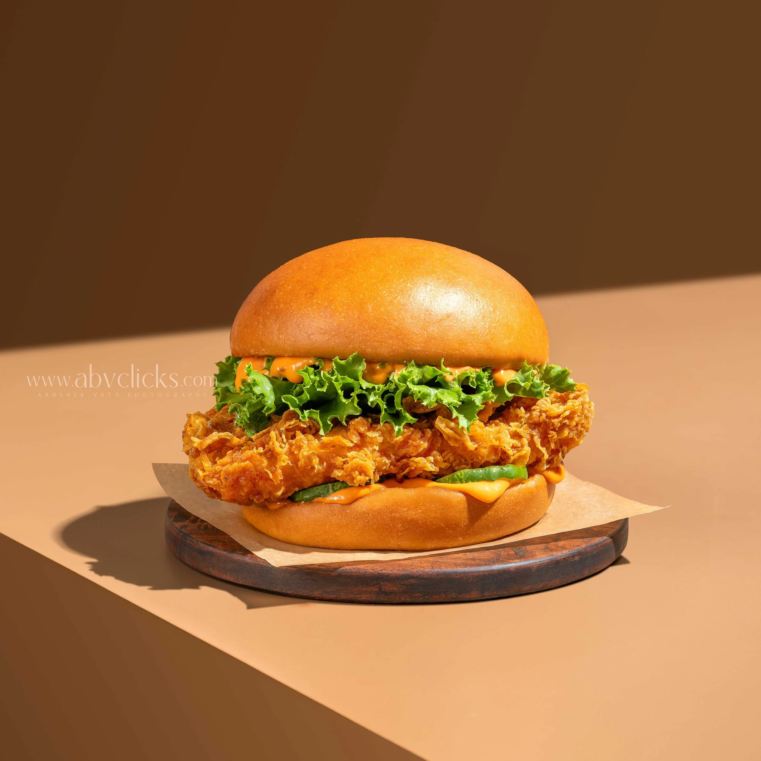 A fried chicken sandwich with leaf lettuce, sliced pickles, and spicy orange sauce in a sandwich bun placed on a wooden board.
