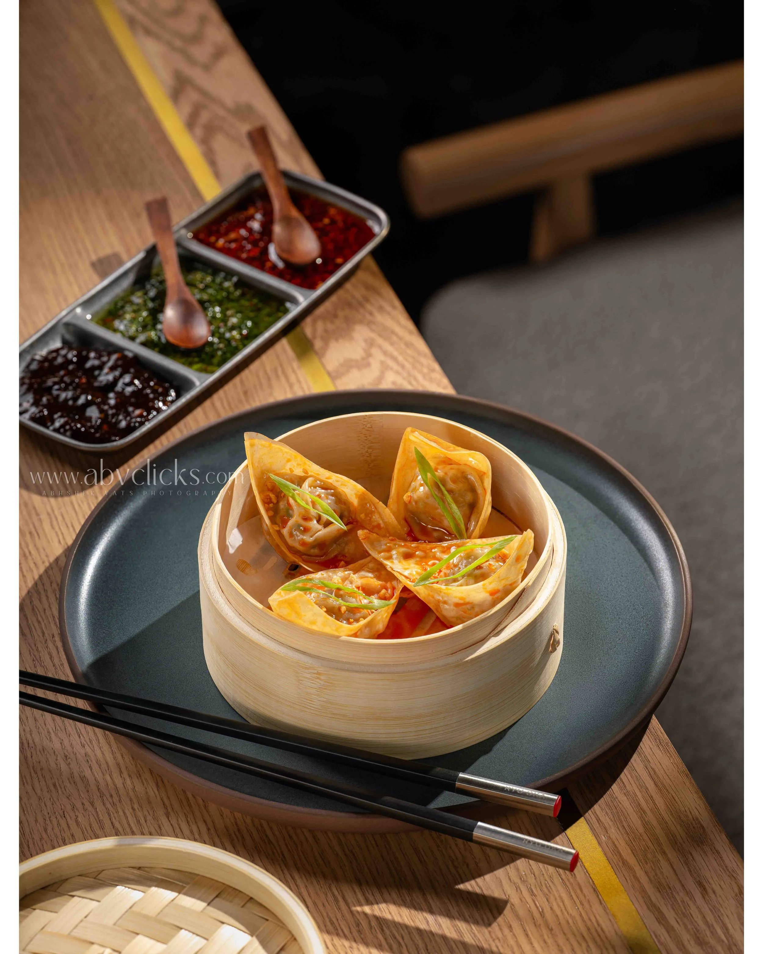 A bamboo steamer basket filled with dumplings on a black plate, placed on a wooden table with chopsticks nearby. In the background, there is a rectangular tray with three sections of dipping sauces, each with a small wooden spoon, on the table.