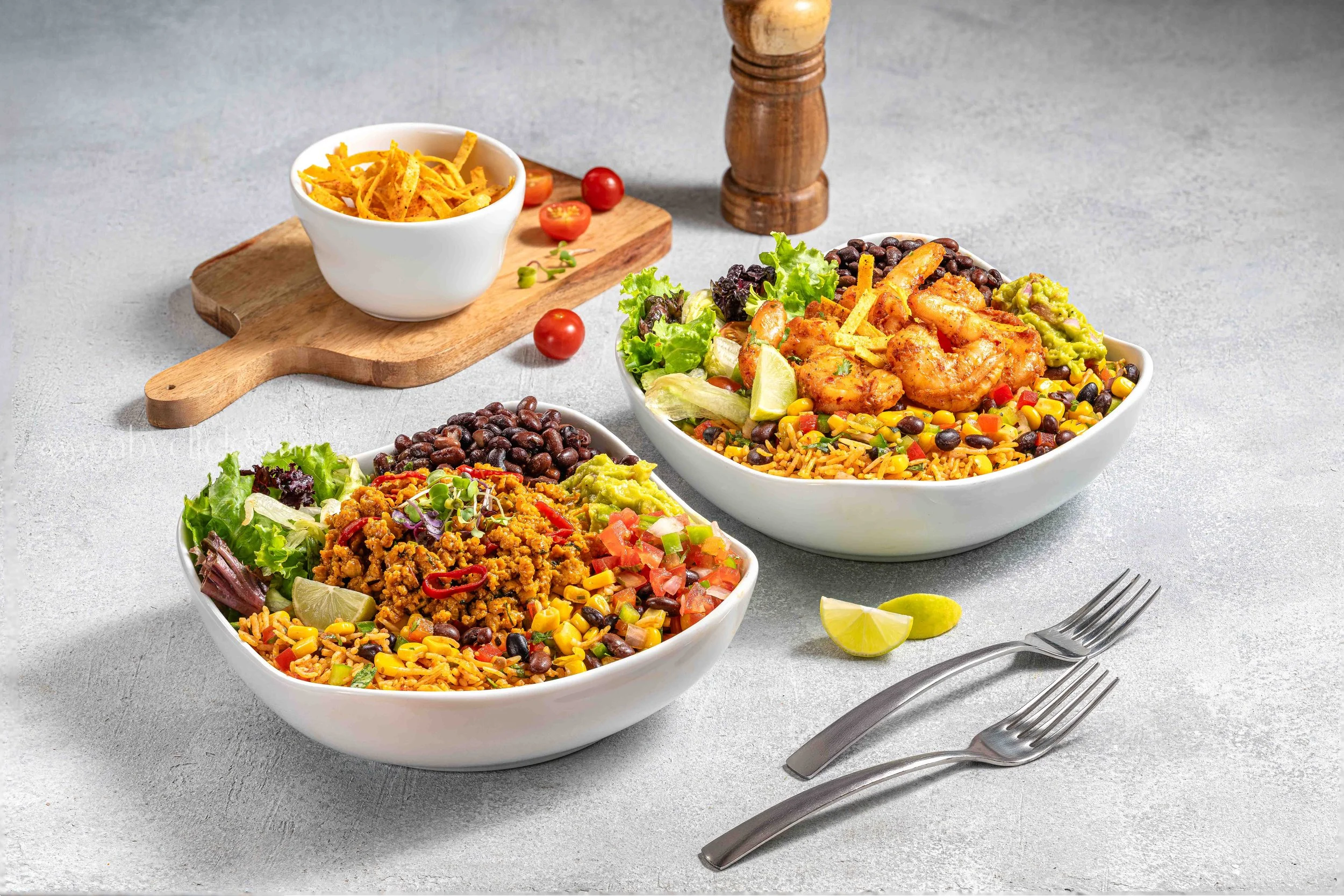 Two large white bowls filled with Mexican-style rice, black beans, lettuce, guacamole, and grilled chicken, garnished with lime wedges, on a gray table with forks nearby, and a wooden pepper grinder in the background.
