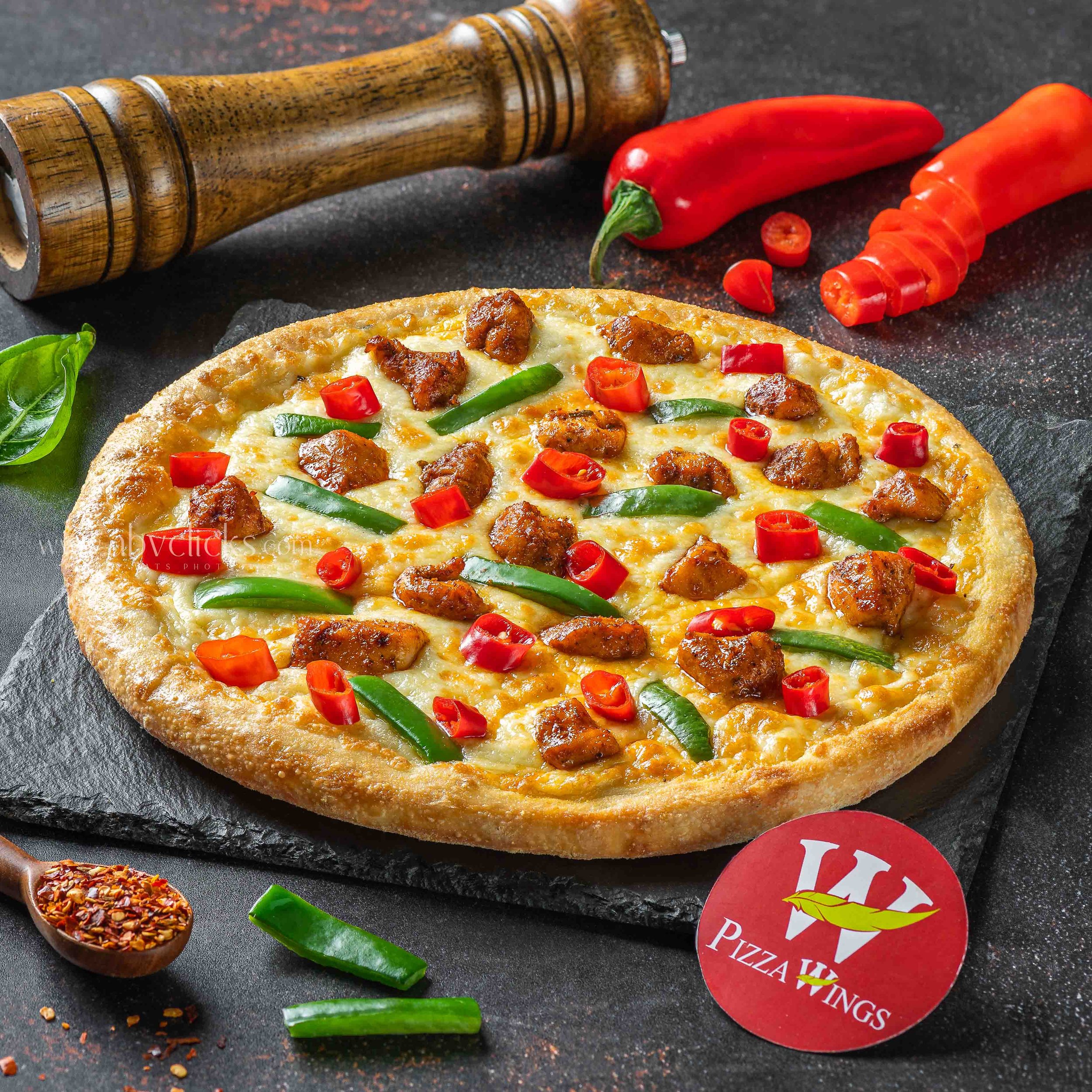 Close-up of a pizza topped with cheese, sausage, and green peppers on a dark surface. Nearby, there is a red pepper, a green pepper sliced, a wooden pepper grinder, and a spoonful of red pepper flakes.