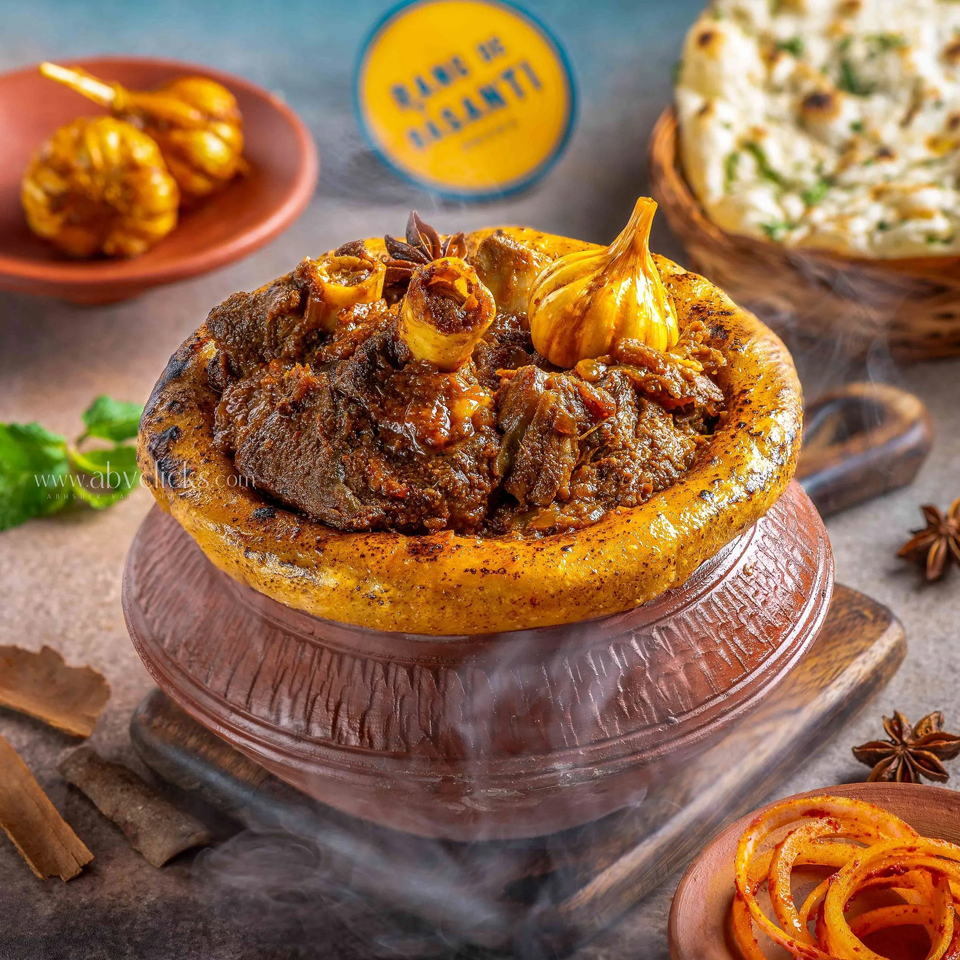A close-up of a traditional Indian dish called galouti kebab served in a hollowed-out bread bun, with additional side dishes including grilled onions, a bowl of salad, and a bowl of seasoned onions with herbs in the background.