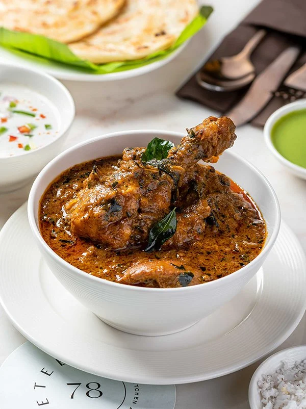 A bowl of Indian chicken curry with a chicken drumstick on top, garnished with curry leaves, served with side dishes including rice, green sauce, and a white yogurt-based dish, all on a white table.