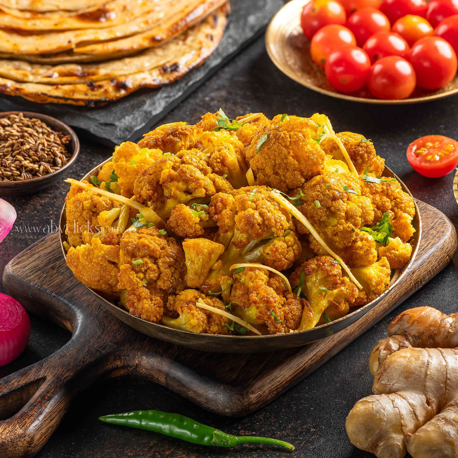 A serving bowl of spicy cauliflower with cilantro garnishing, placed on a wooden serving board, surrounded by cherry tomatoes, radishes, roasted chickpeas, green chili, flatbread, and other ingredients on a dark surface.