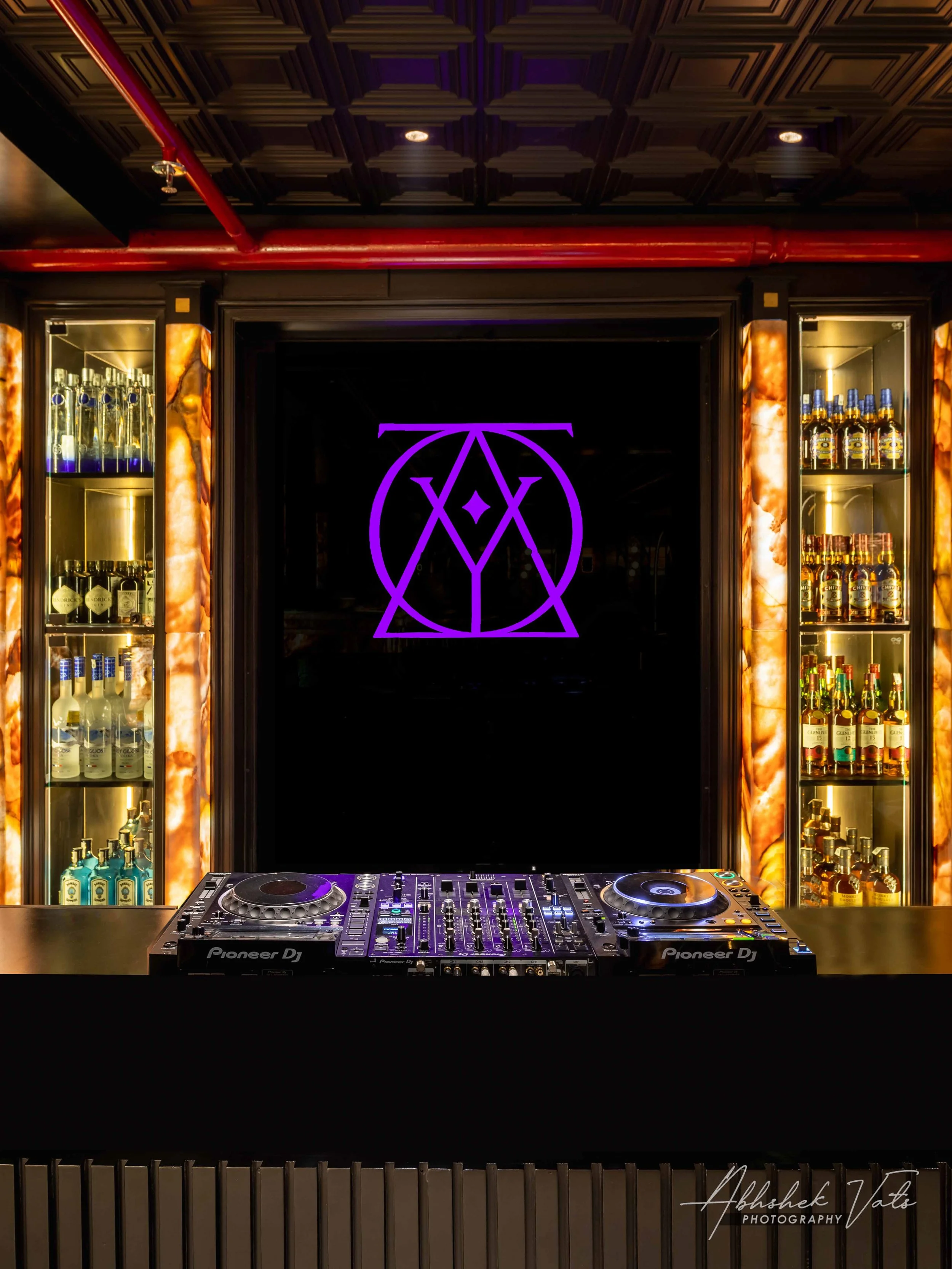 A DJ setup with Pioneer DJ controller in front of illuminated shelves with bottles of spirits and liquors, and a neon purple symbol on a black screen behind. The ceiling has a geometric wood pattern and red pipes running across.