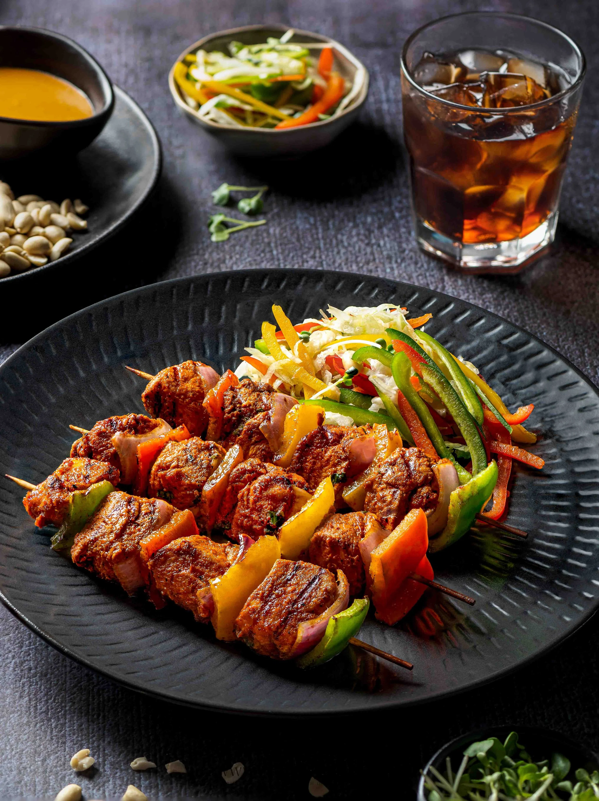 A black plate of grilled chicken skewers with colorful bell peppers and onions, served with a side of fresh vegetable salad and a glass of iced cola.