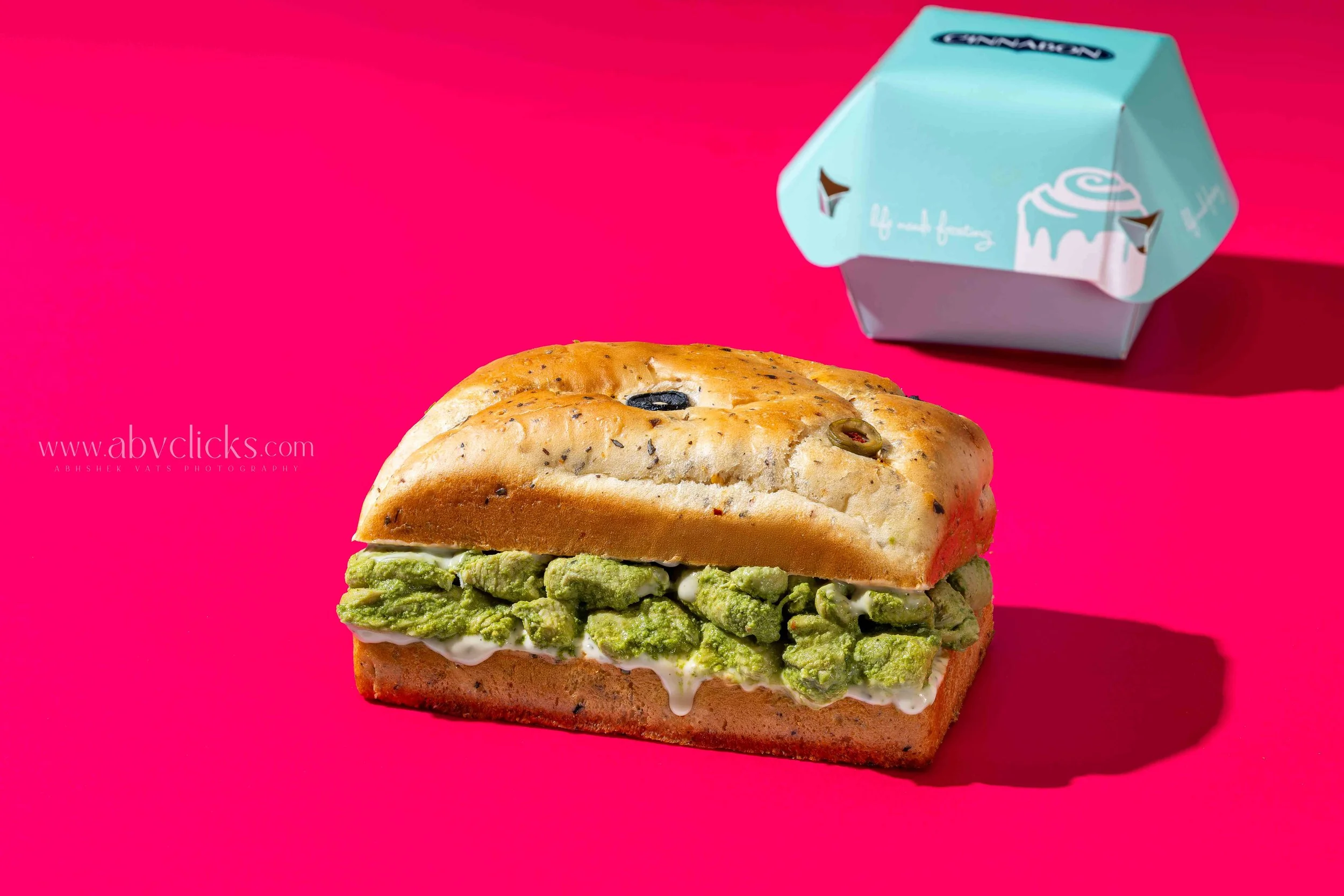 Close-up of a sandwich with green pesto, white sauce, and black olives on toasted bread, on a bright pink background with a light blue boxed cupcake in the upper right corner.