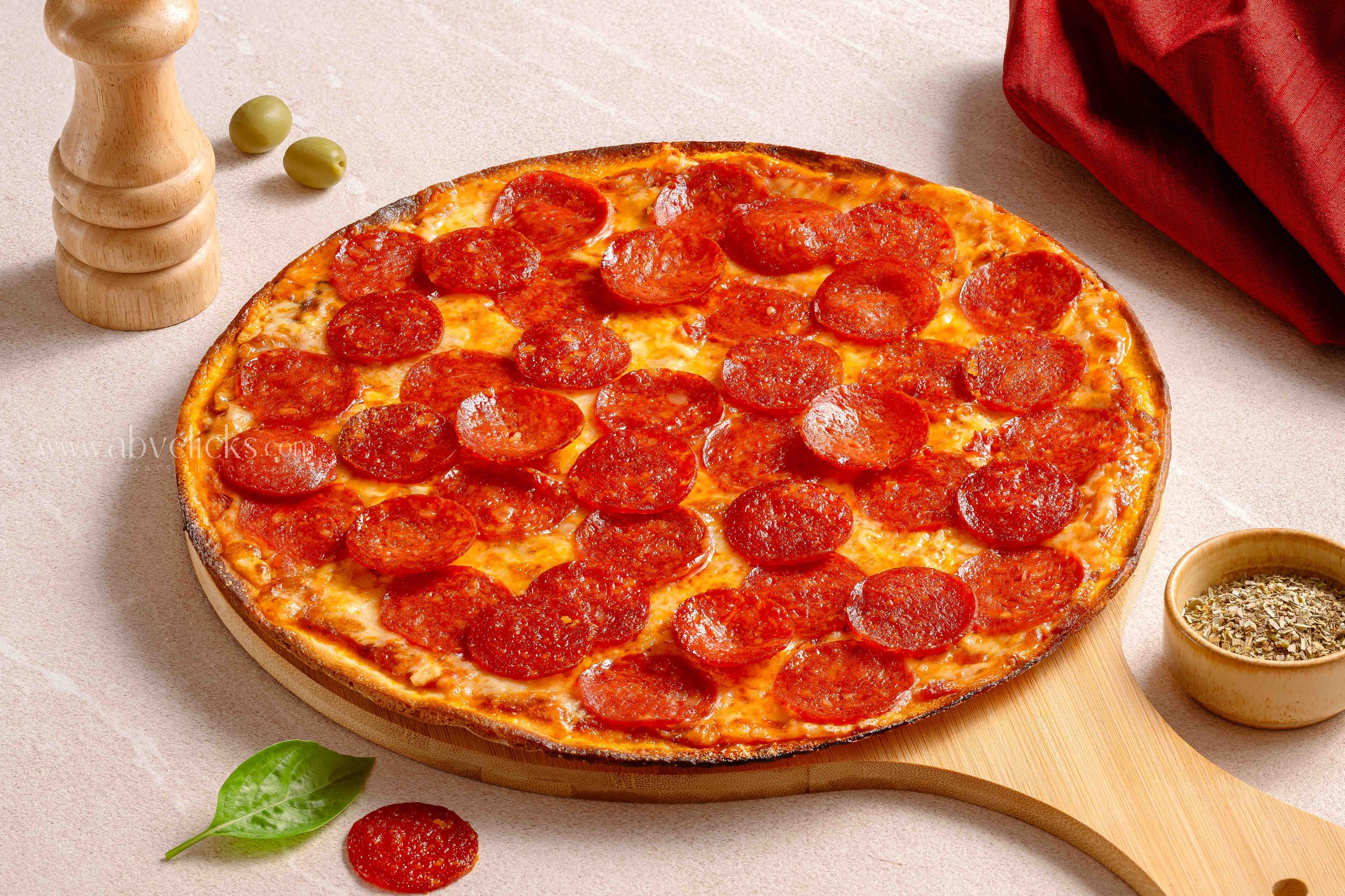 Pepperoni pizza on a round wooden board with a red cloth, green olives, and spices nearby.
