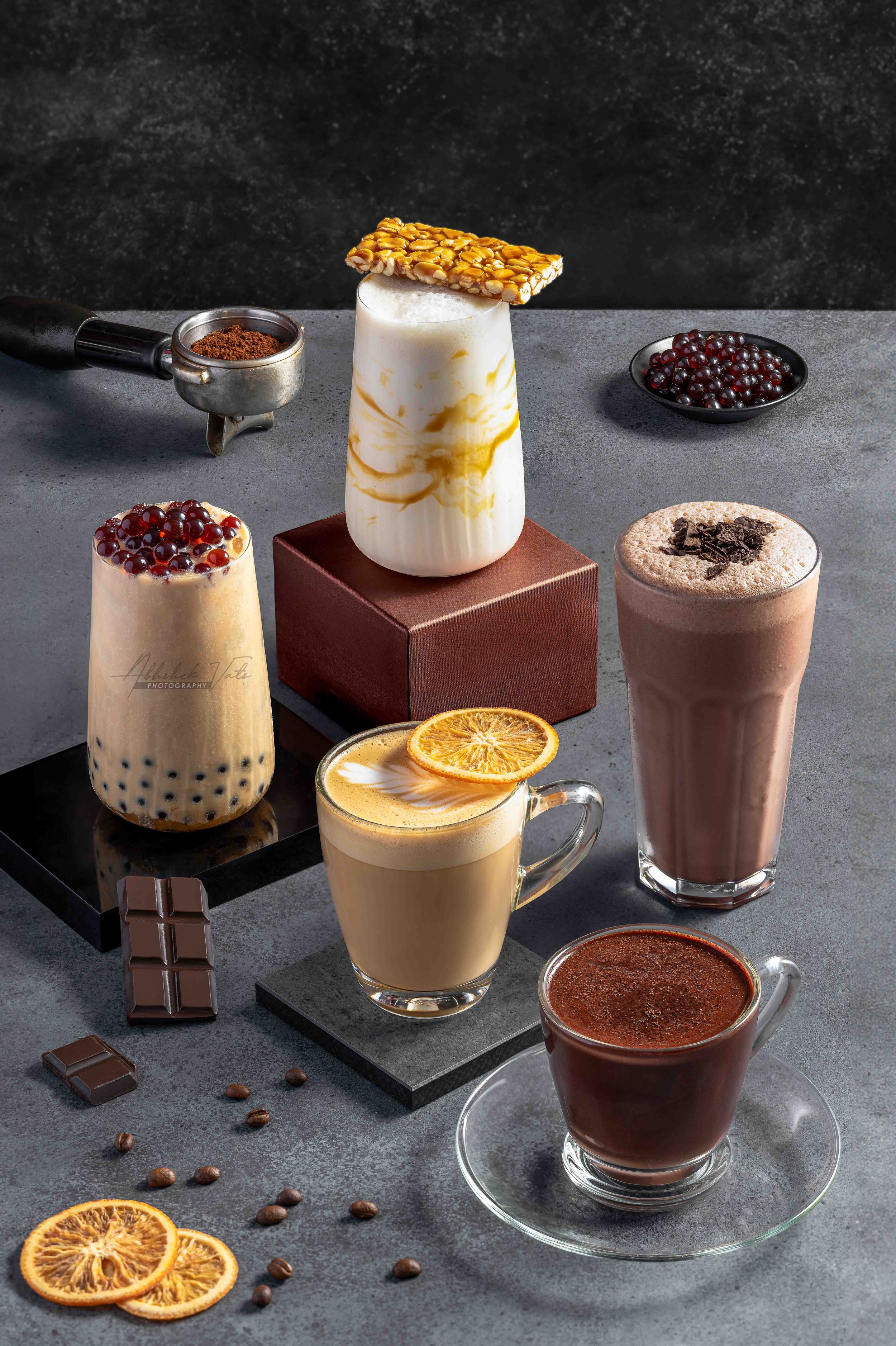 Various coffee drinks on a dark gray surface, including a milkshake with boba pearls, a frothy coffee topped with orange slices, a hot chocolate with cinnamon, and other beverages with whipped cream and toppings. There are also chocolate bars, cocoa 