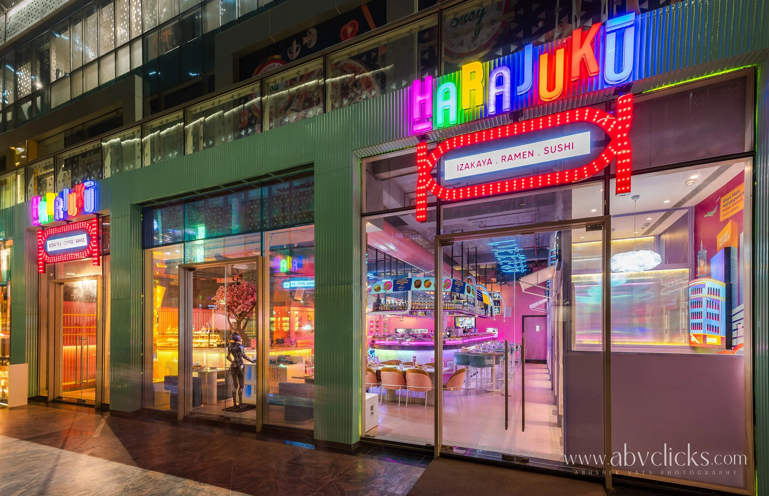 Colors neon sign reading 'HARAJUAN,' above a restaurant entrance with neon signs indicating 'IZAKAYA, RAMEN, SUSHI.' The restaurant has glass walls revealing a brightly lit interior with pink and yellow lighting, beige chairs, and decorative elements