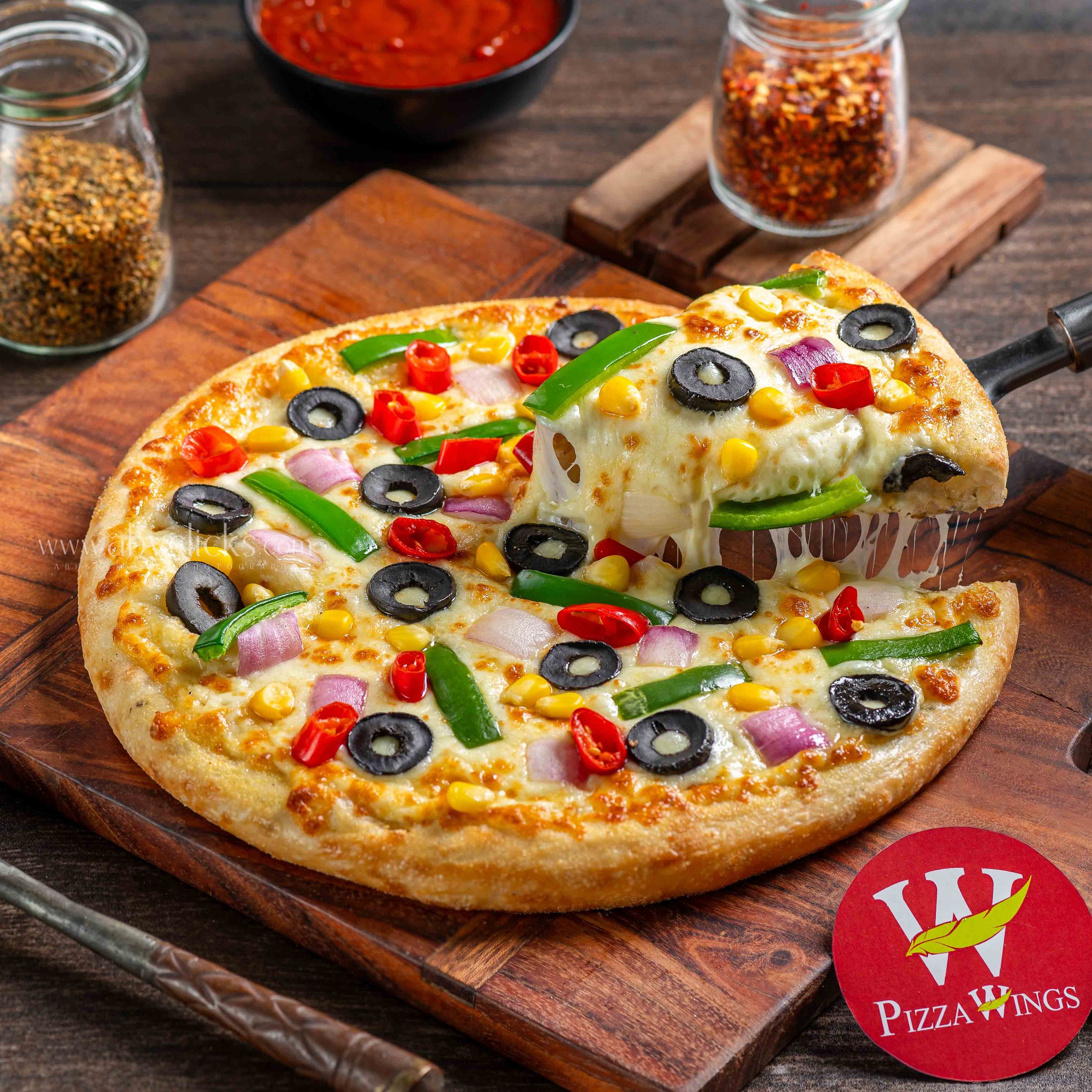 Close-up of a cheese pizza topped with black olives, green peppers, red peppers, yellow corn, and red onions on a wooden cutting board, with a jar of red pepper flakes, a jar of mixed spices, and a bowl of marinara sauce in the background.