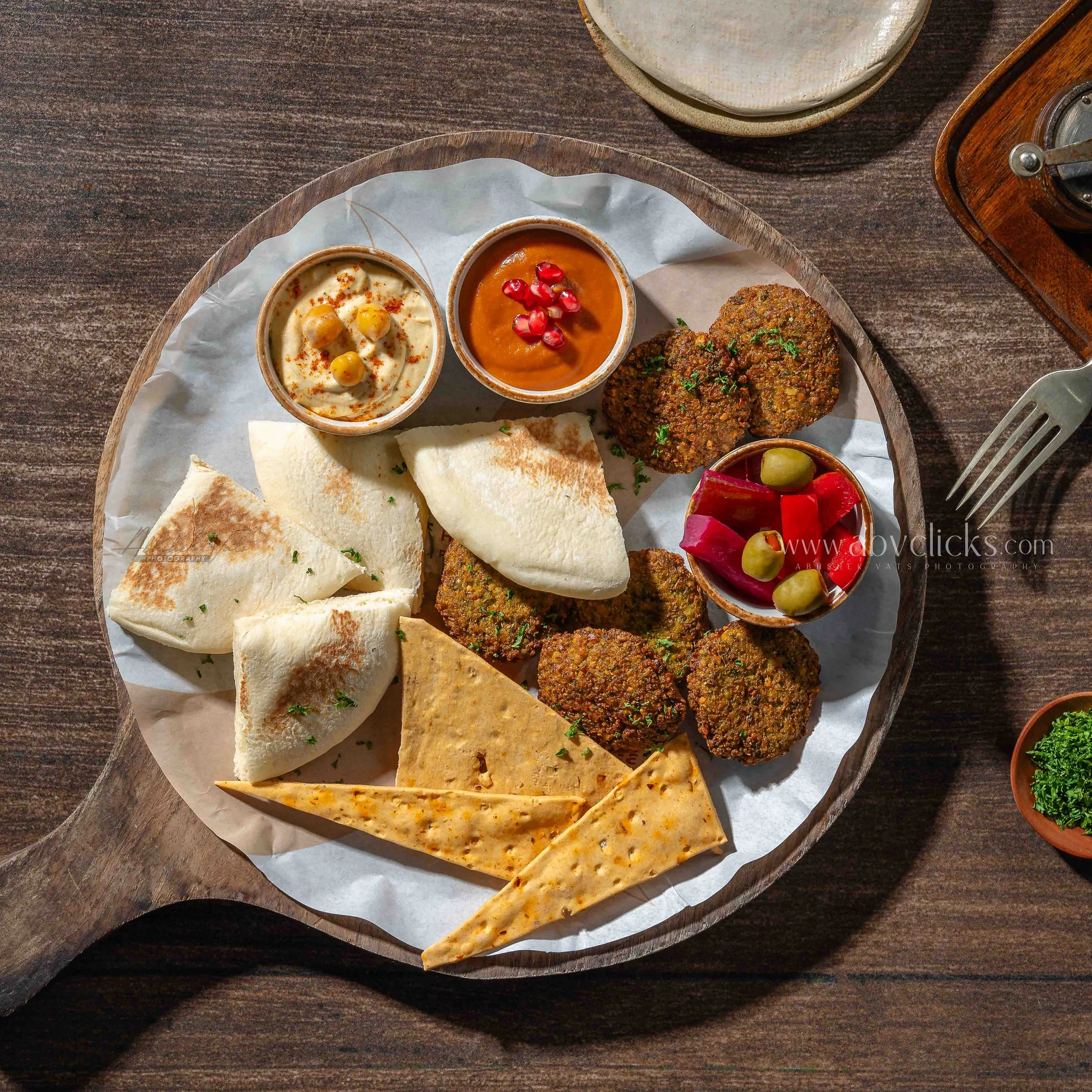 A round wooden tray with various Indian dishes including naan bread, chickpea curry, a tomato-based curry with pomegranate seeds, and falafel with salad on the side, placed on a dark wooden table.