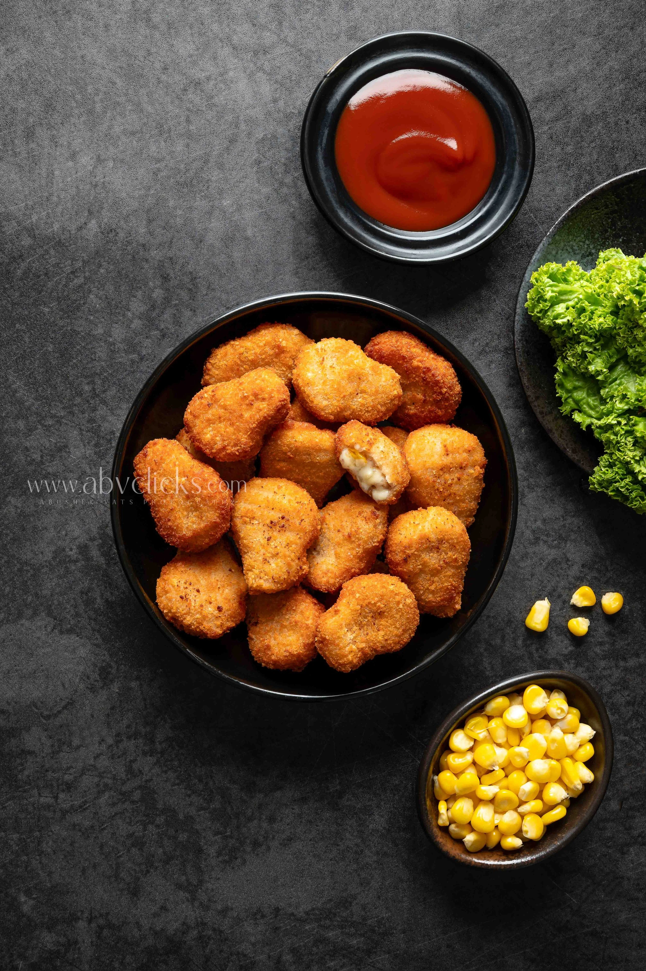 A black bowl filled with breaded chicken nuggets on a dark surface. To the right, a black bowl with yellow corn kernels. In the top right corner, part of a black plate with fresh green lettuce. Near the chicken nuggets, there is a small black bowl of