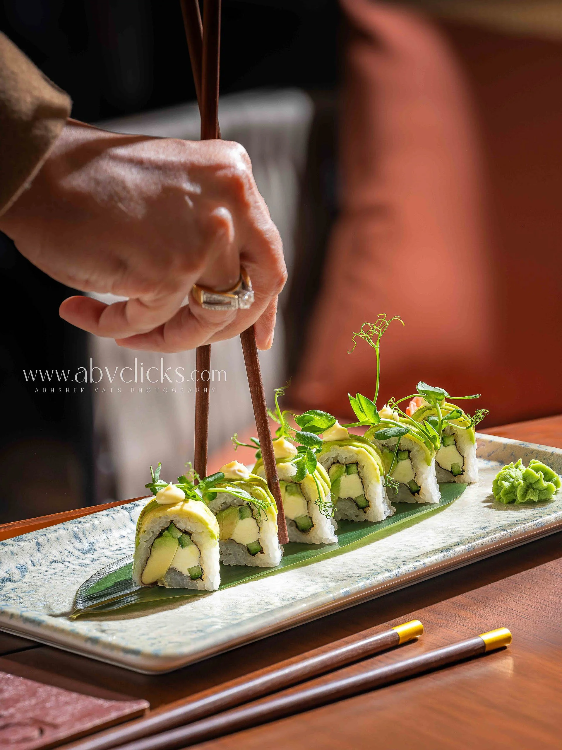 Close-up of a person's hands using chopsticks to pick up a piece of sushi on a rectangular ceramic plate, with colorful sushi rolls garnished with greens and wasabi.