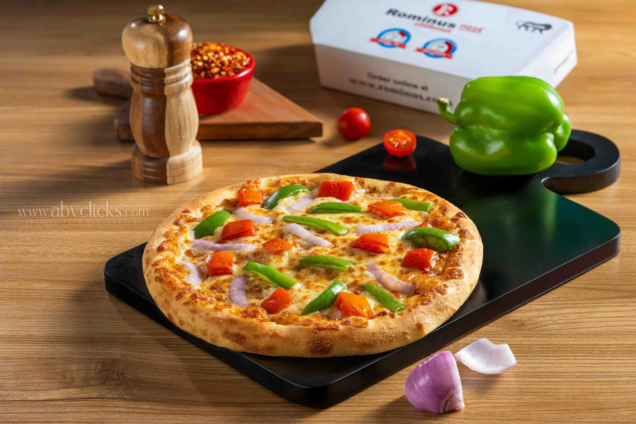 A freshly baked pizza with green bell peppers, red bell peppers, and onions on a black cutting board. In the background, there is a green bell pepper, cherry tomatoes, a container of crushed red pepper, and a pizza box on a wooden table.