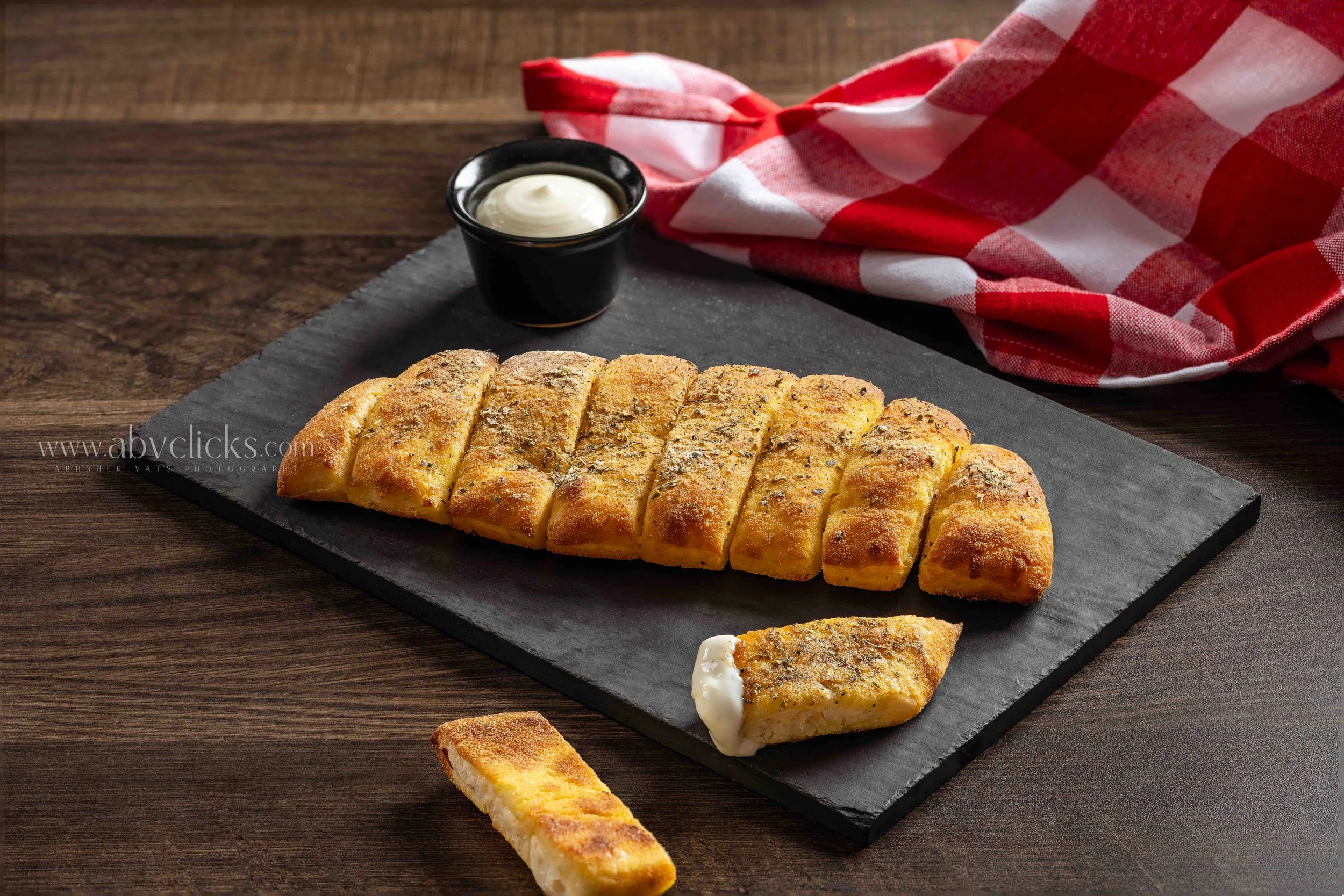 Slices of garlic bread on a black slate serving board with a small black dish of creamy dip and a red and white checkered napkin in the background.