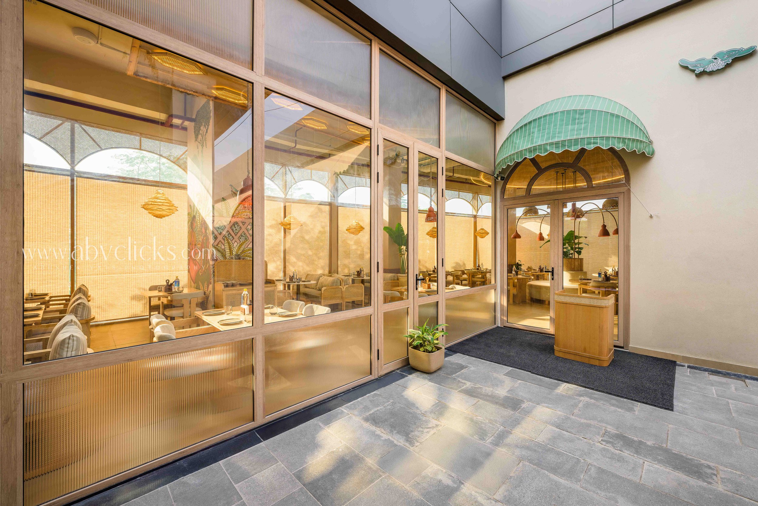 Exterior view of a modern restaurant with large glass windows and a green striped awning. The interior, visible through the glass, shows tables, chairs, and hanging lights. The pavement outside is made of gray stone tiles.