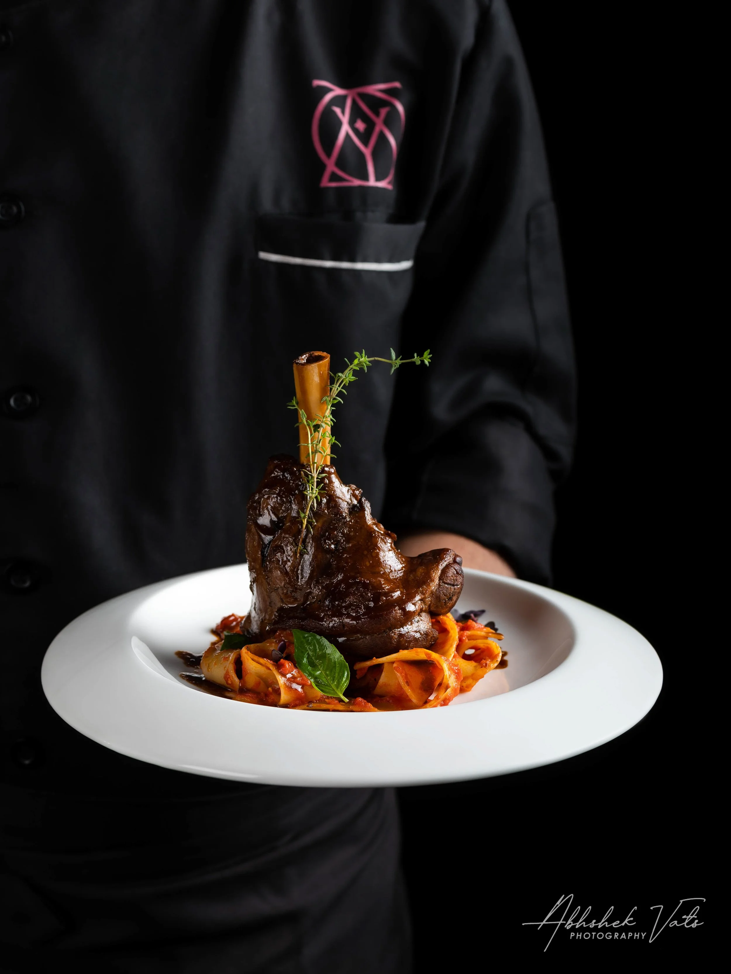 A chef dressed in black holding a white plate with a cooked lamb shank garnished with a sprig of thyme, served over a bed of pasta with sauce and basil leaves.