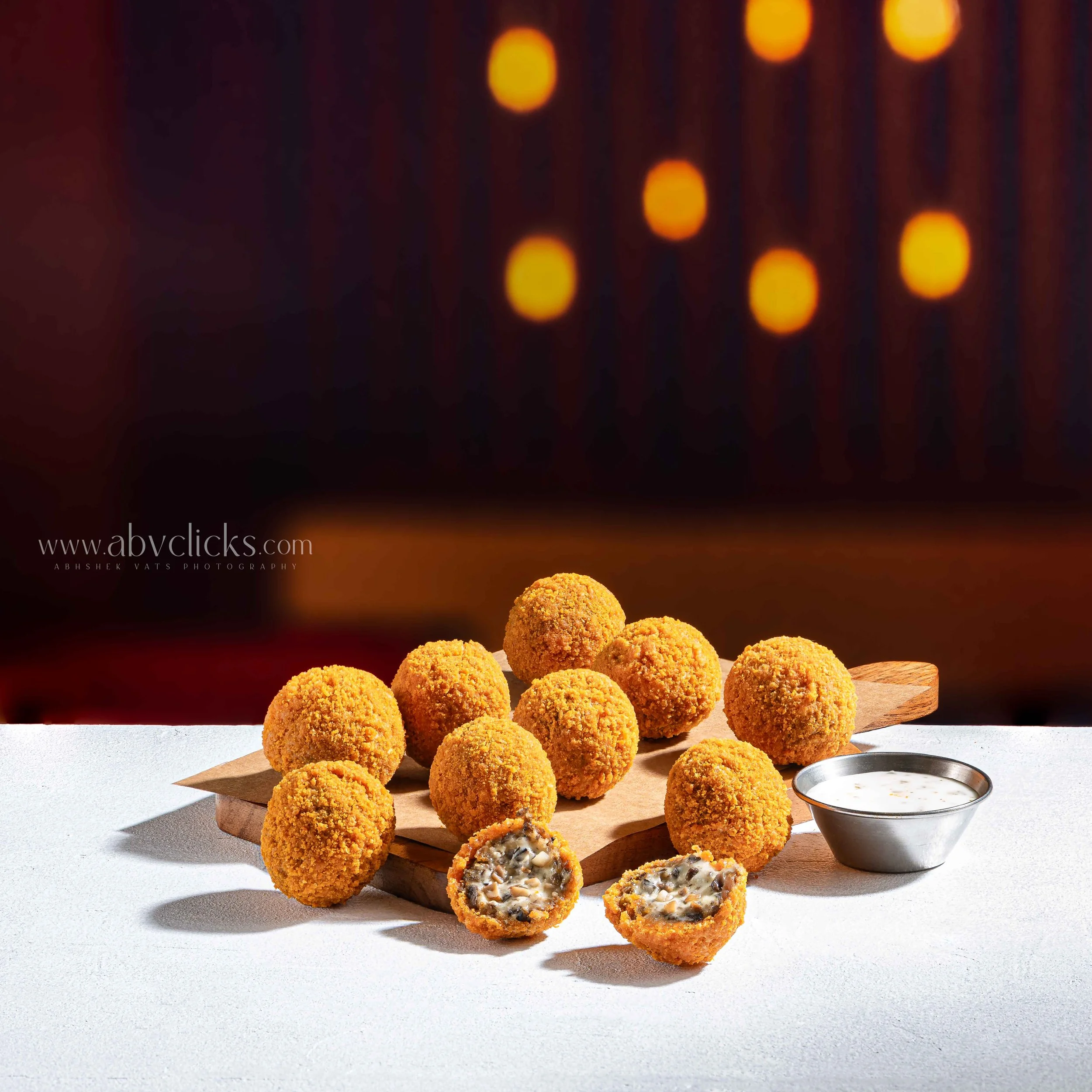 A pile of breaded and fried bunches of white and black sesame seed sweet or savory balls on a wooden cutting board, with one broken open showing a creamy black and white filling, next to a small metal dish of white dipping sauce.