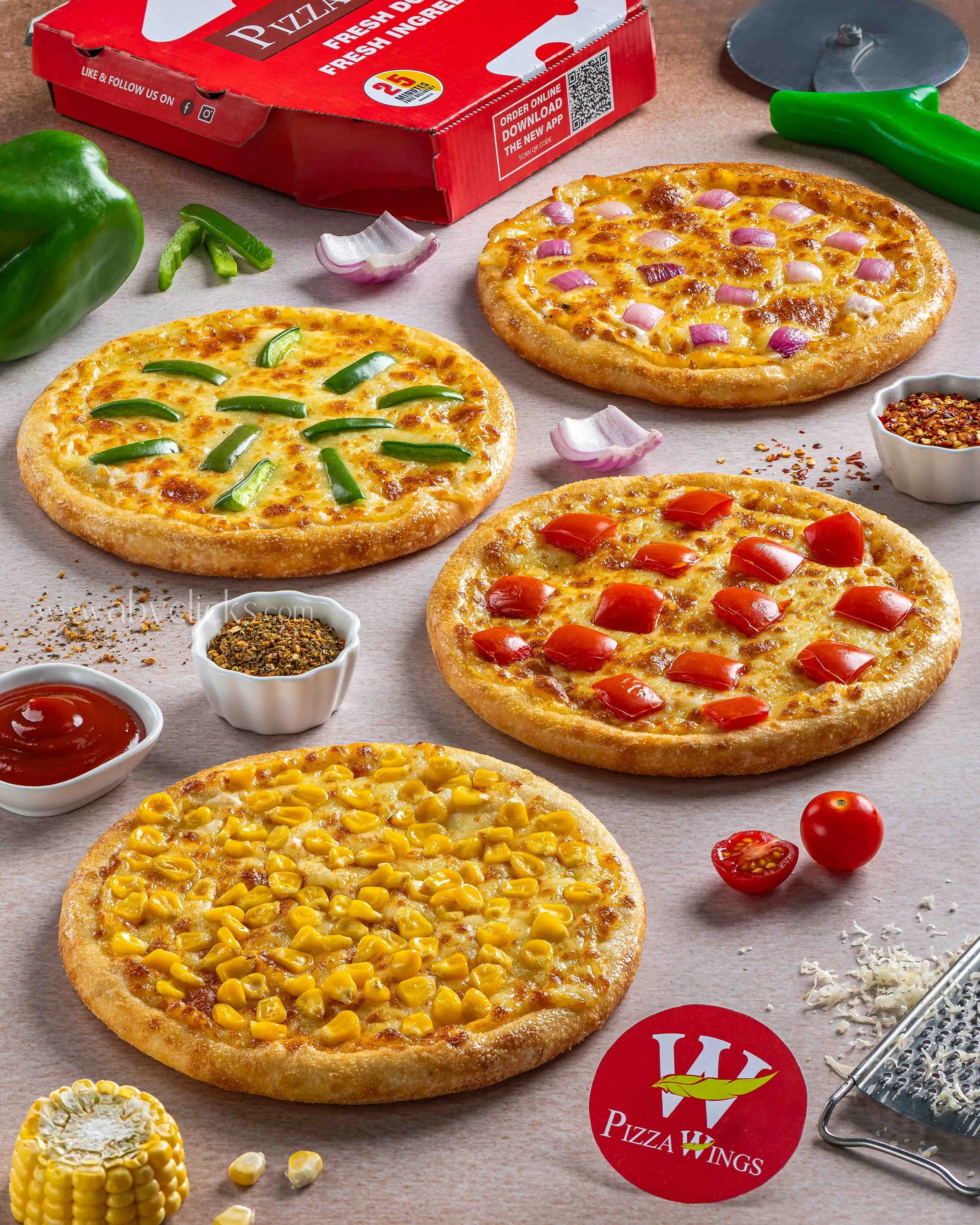 Three pizzas with cheese and different toppings on a wooden surface, surrounded by a green bell pepper, cherry tomatoes, small bowls of spices, and kitchen tools, with a red pizza box in the background.