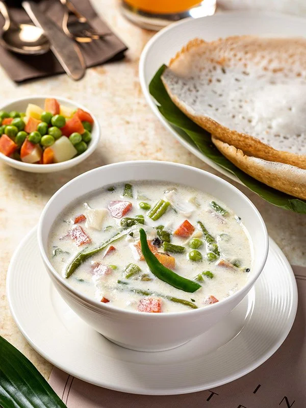 A bowl of Thai green curry with chicken and vegetables, a side of colorful mixed vegetables, and an empty papadam on a plate.