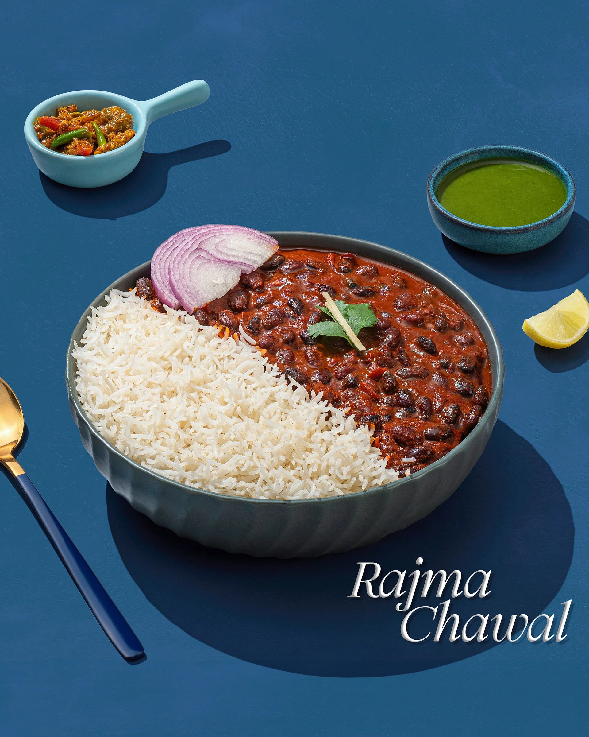 A bowl of Indian chana chaat, orange-red with black chickpeas, sliced red onion, rice, lemon wedge, and cilantro, with a small bowl of curry and a green chutney on a blue surface.