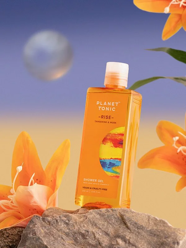 A bottle of Planet Tonic Rise Shower Gel with tangerine and musk scent, placed on rocks surrounded by orange lilies and flowers, with a blurred planetary object in the background and a colorful, nature-inspired label.