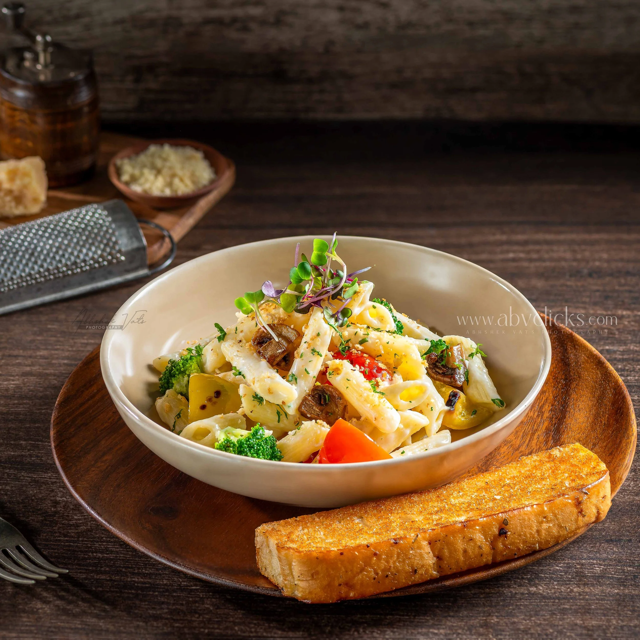 A bowl of creamy pasta topped with microgreens on a wooden plate, with bread and grated cheese in the background.