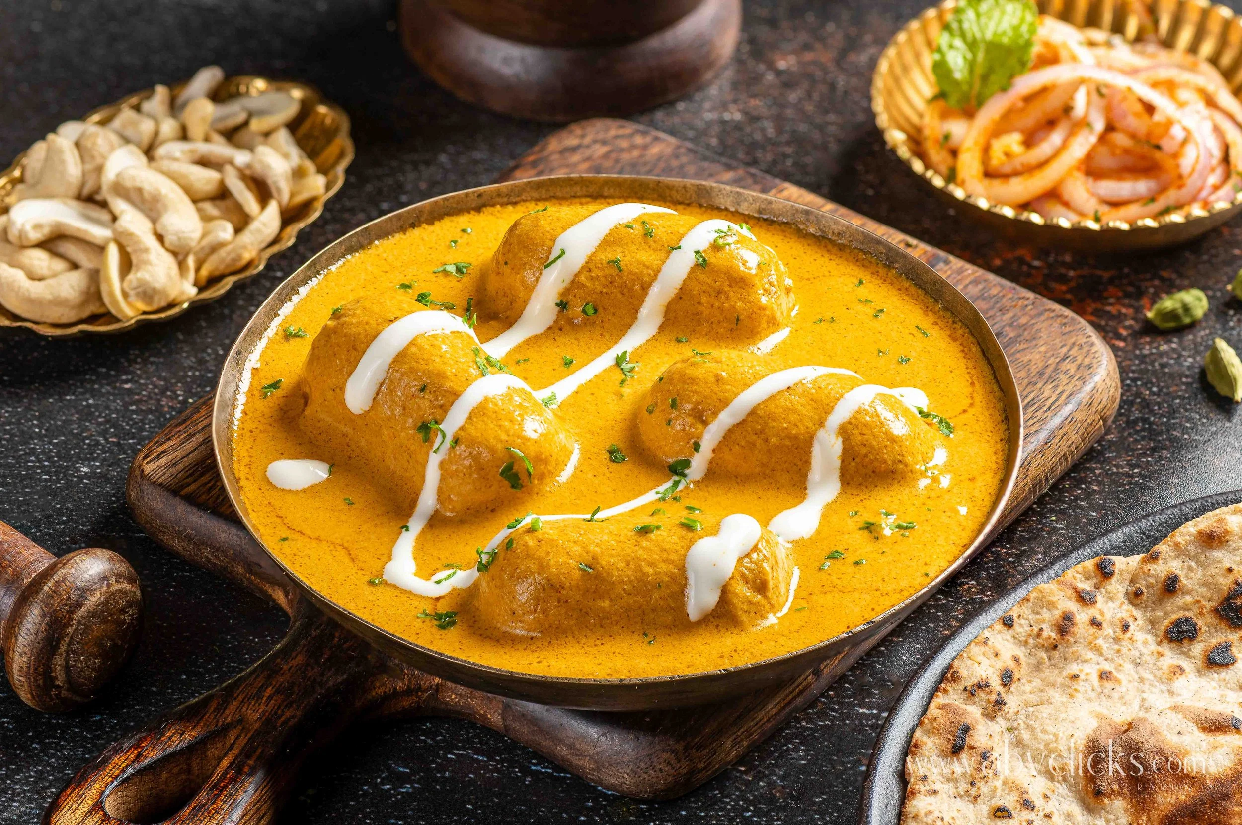 A close-up of a bowl of chicken curry with a creamy yellow sauce, garnished with white cream and chopped herbs, on a wooden serving board. Surrounding dishes include a bowl of cashew nuts, a bowl of salad with sliced onions, green herbs, and a piece 