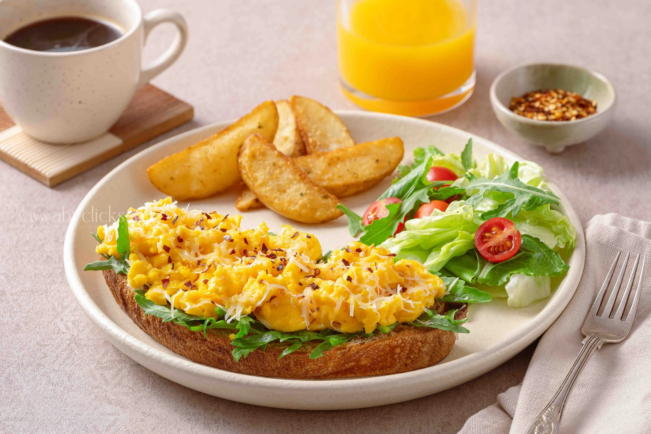 Open-faced egg salad sandwich on whole grain bread with arugula and cherry tomatoes, served with potato wedges, a side salad, a glass of orange juice, a cup of coffee, and a bowl of chili flakes.