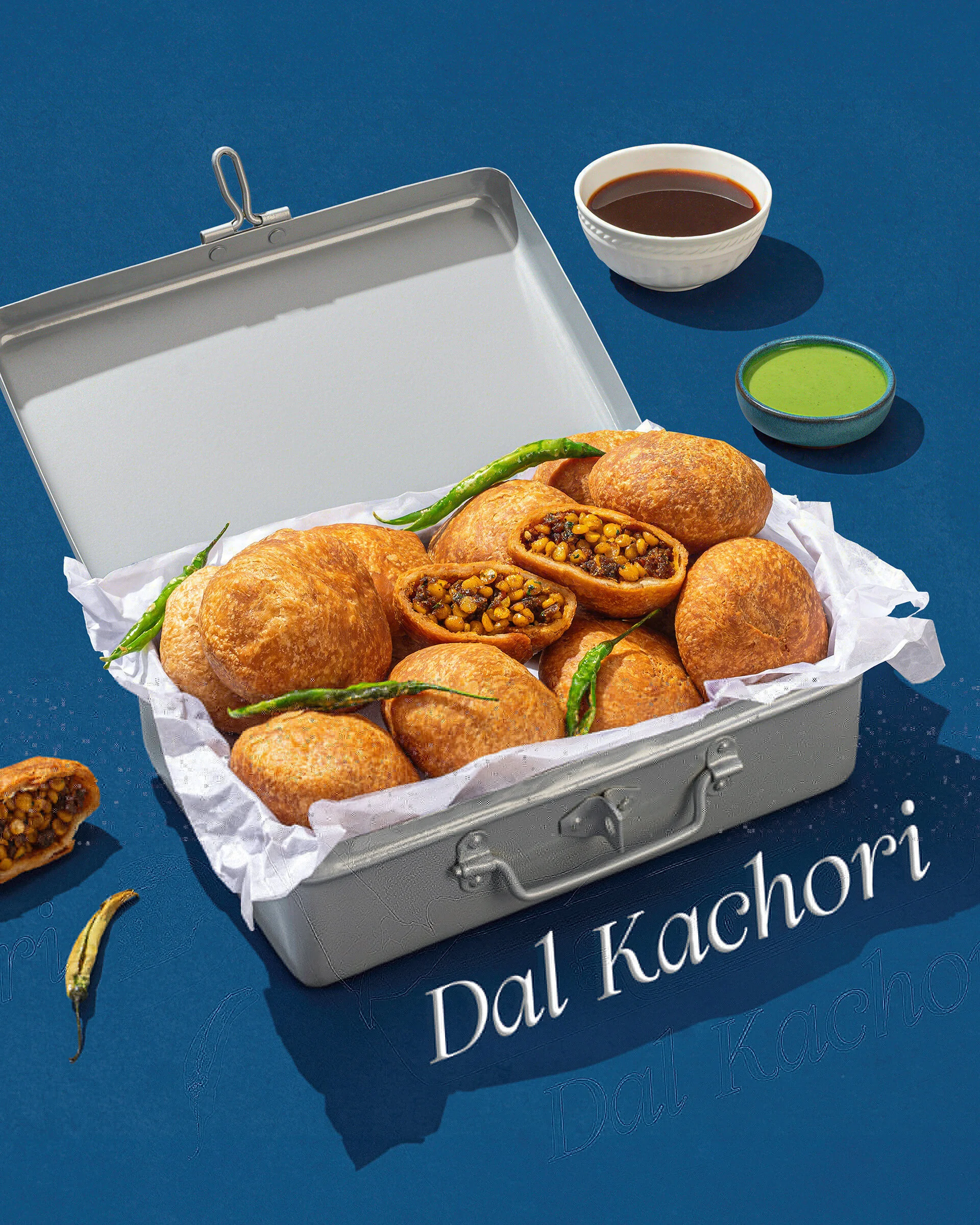 Metal container with parchment paper holding fried pastries filled with a bean mixture, green chili peppers, and two bowls of sauces, one dark and one green, on a blue surface with text partially visible.