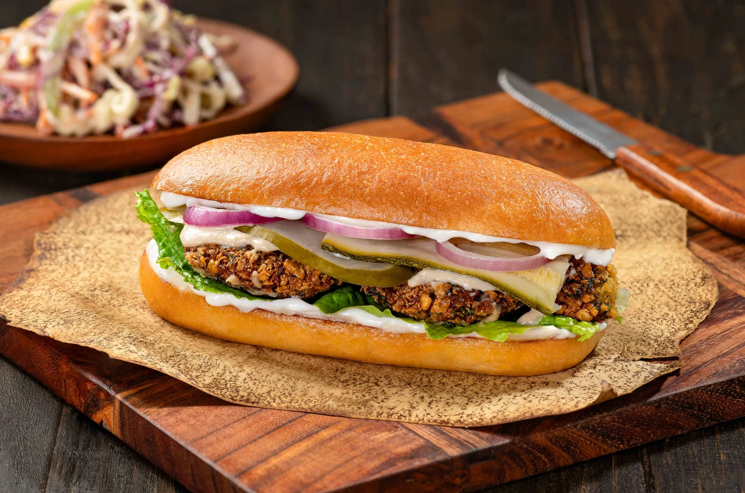 A sandwich with a bread roll filled with fried meat, lettuce, pickles, onions, and mayonnaise, placed on a wooden board with a side of coleslaw in the background.