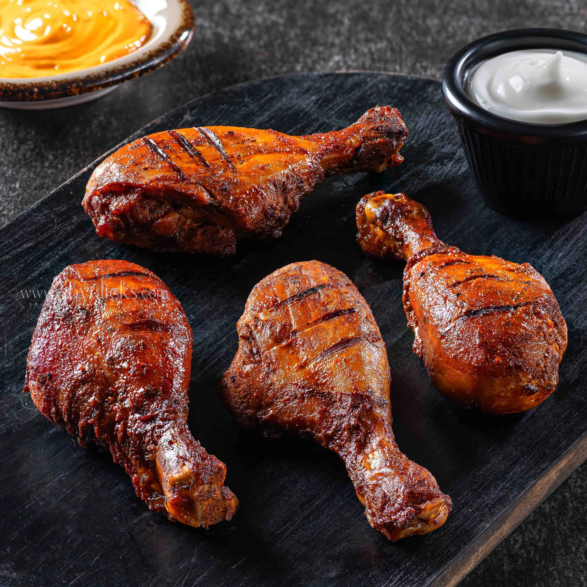 Close-up of grilled pork ribs with a glaze, served on a black wooden board, with small bowls of orange sauce and white dipping sauce in the background.