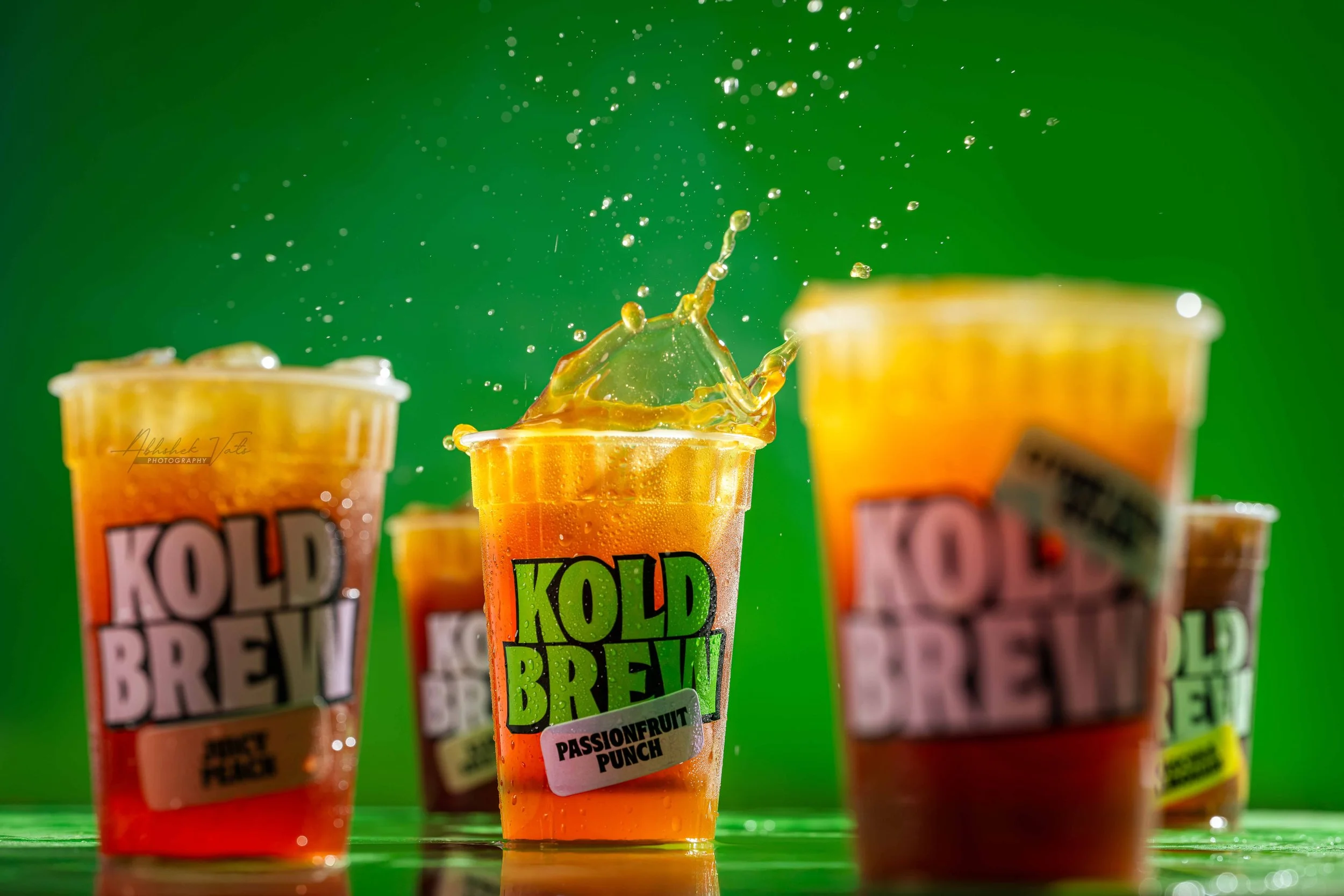 Three bottles of cold beverage labeled 'Kold Brew' with one bottle in the foreground showing a splash of liquid and fruit flavor 'Passionfruit Punch'. The background is green with a blurred effect.