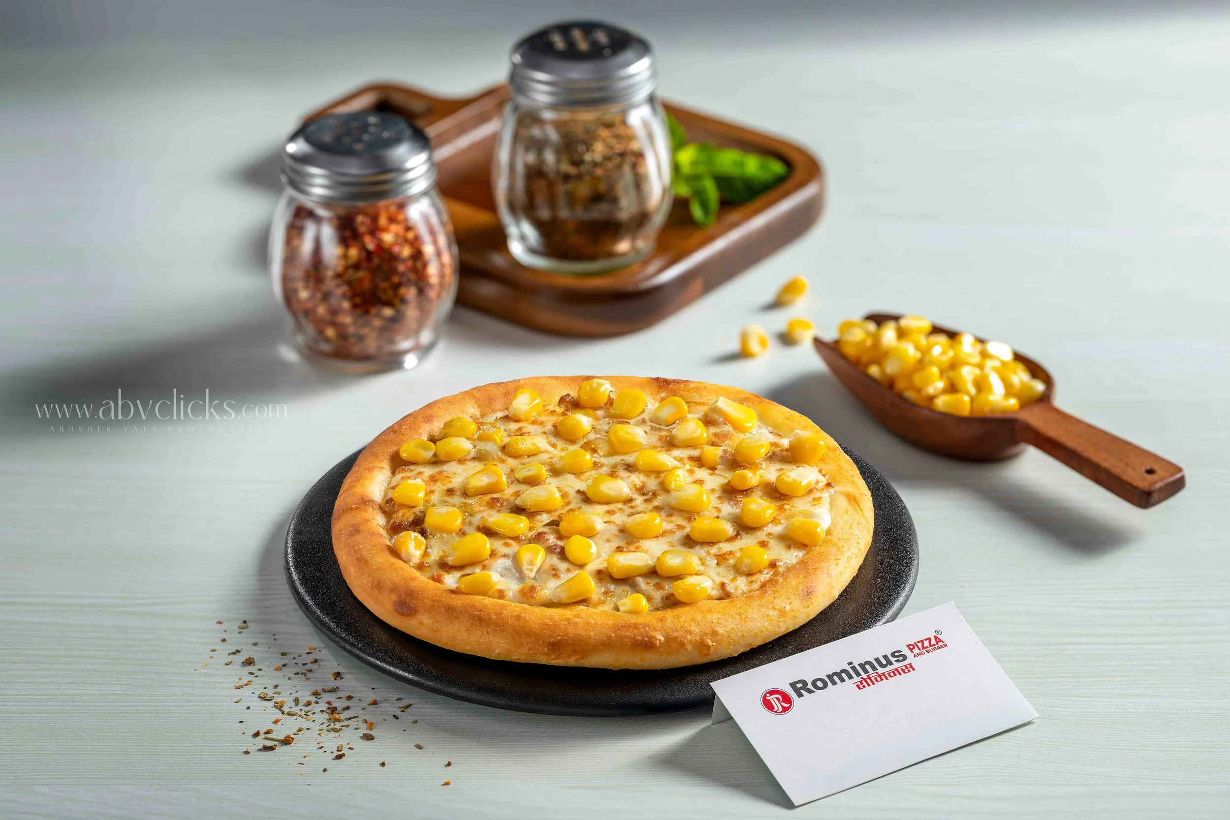 A pizza with corn topping on a black plate, with pepper flakes and seasonings scattered around. In the background, there are two spice shakers, a wooden tray with green herbs, a wooden dish with additional corn, and a piece of paper with the logo 'Ro